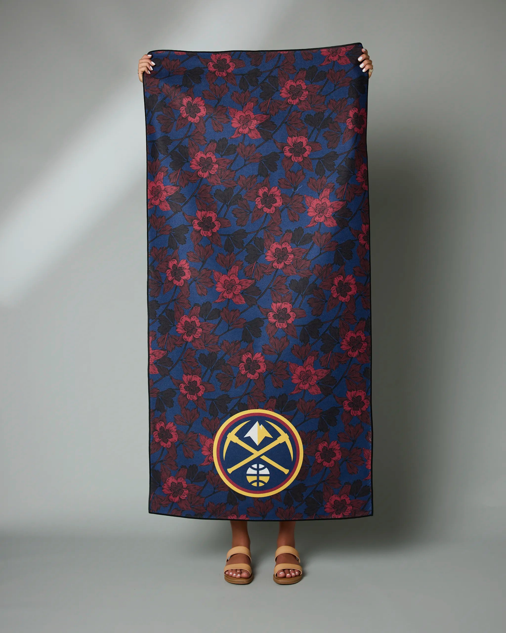 A person holds the Geometry Nuggets Floral towel, featuring a rectangular shape with dark blue and red flowers and the Denver Nuggets logo at the bottom. Only their hands, feet, and sandals are visible.