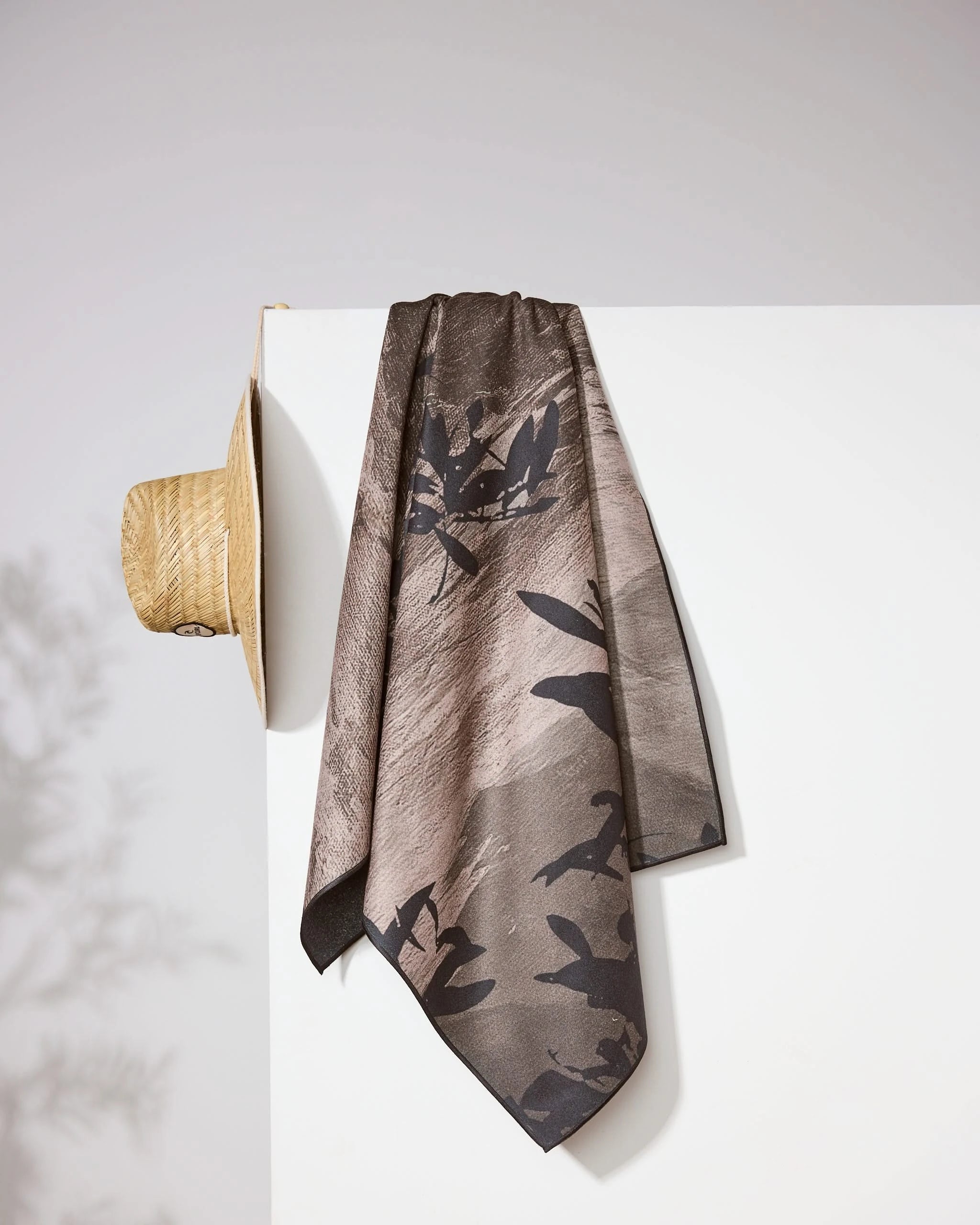 A wide-brimmed straw hat and a patterned brown and black scarf hang on the corner of a white wall, with soft shadows in the background, alongside the Geometry Oaklen Micro-loop Beach Towel made from recycled materials.