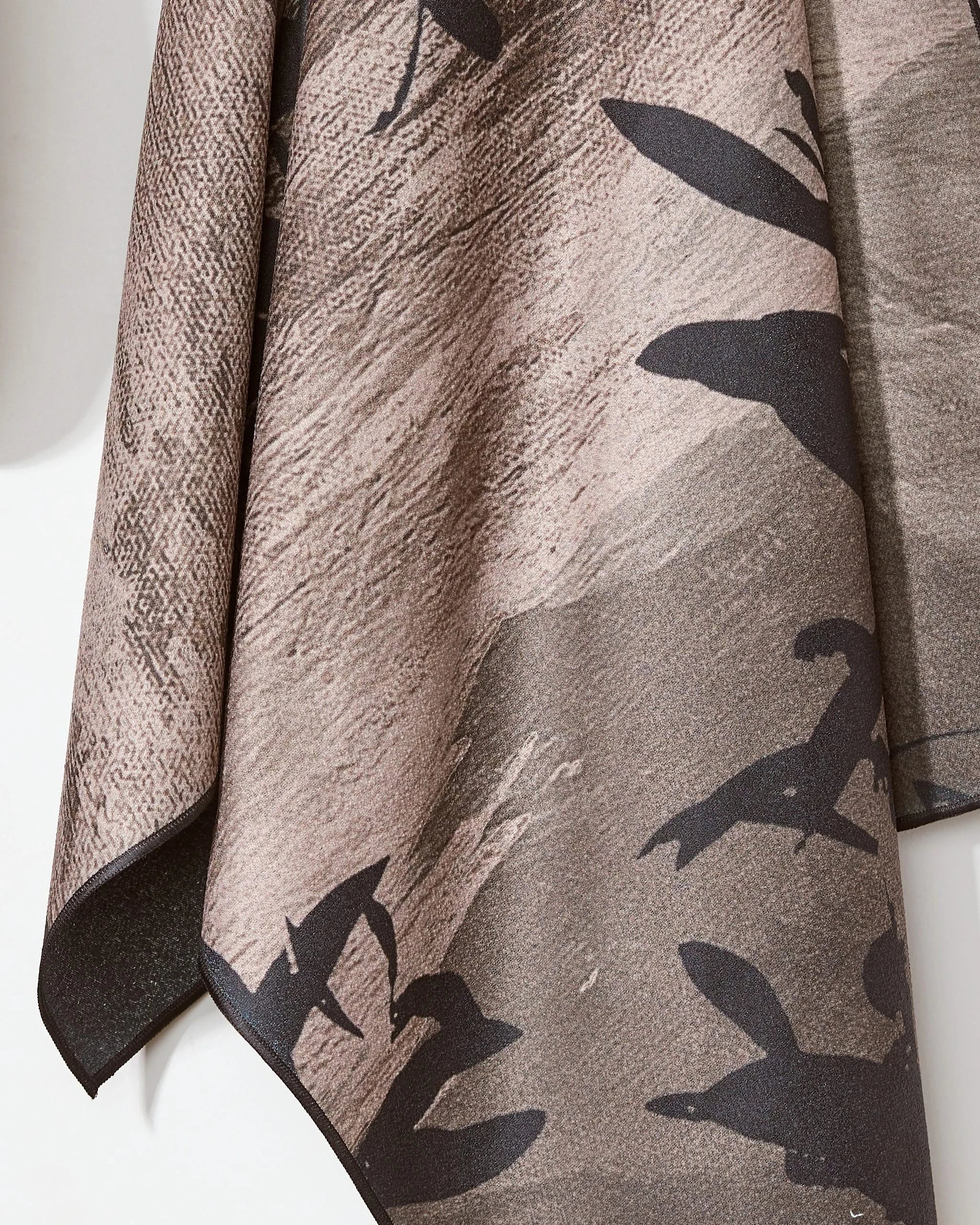 A close-up of the Oaklen Beach Towel by Geometry, hanging against an earthy brown background with black abstract animal-like shapes. The soft, draped material looks ideal for relaxing on the beach.