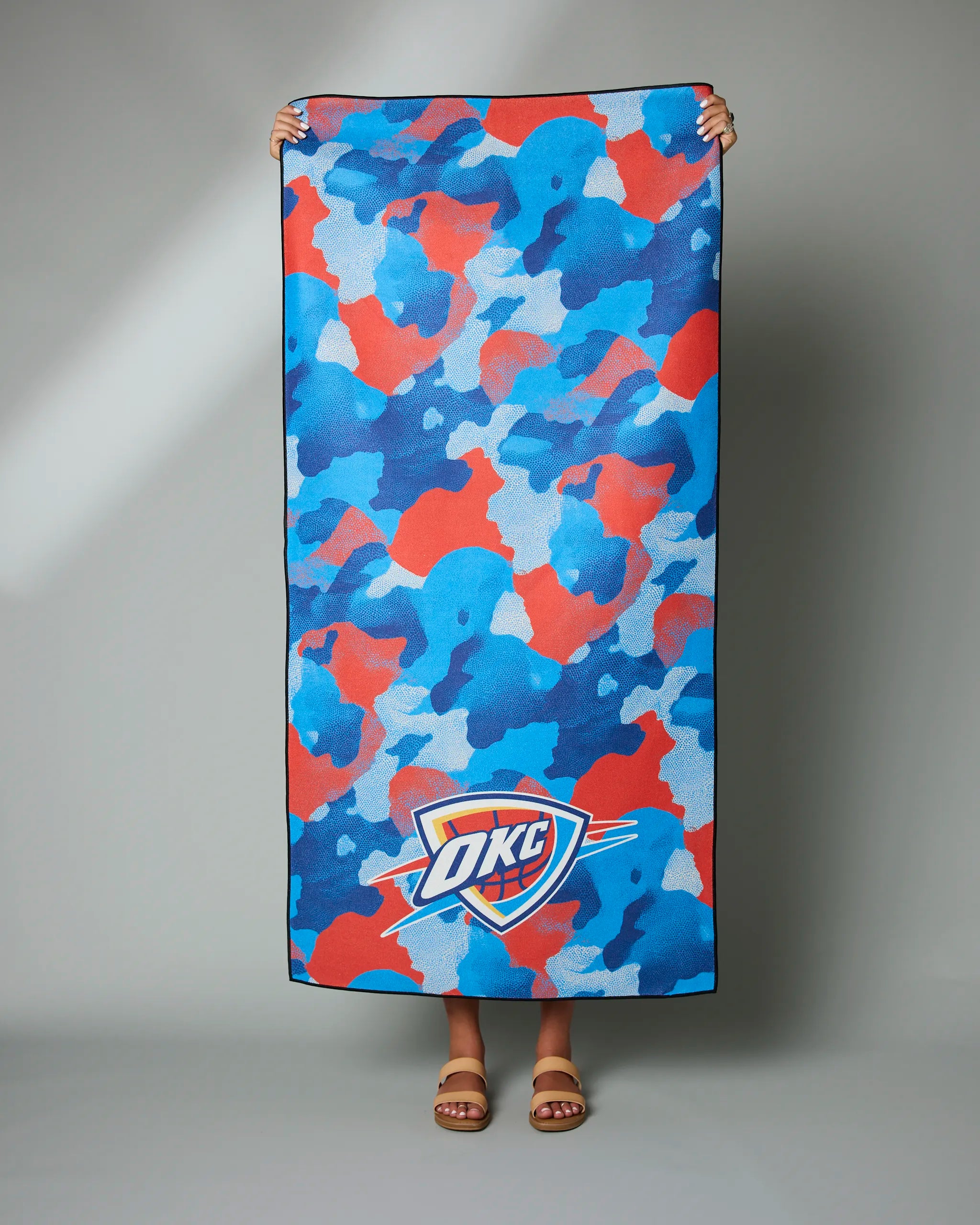 A person holds up the Geometry OKC Camo towel featuring a blue, red, and light blue camouflage design and the OKC Thunder logo at the bottom. Only their hands, feet, and sandals are visible.