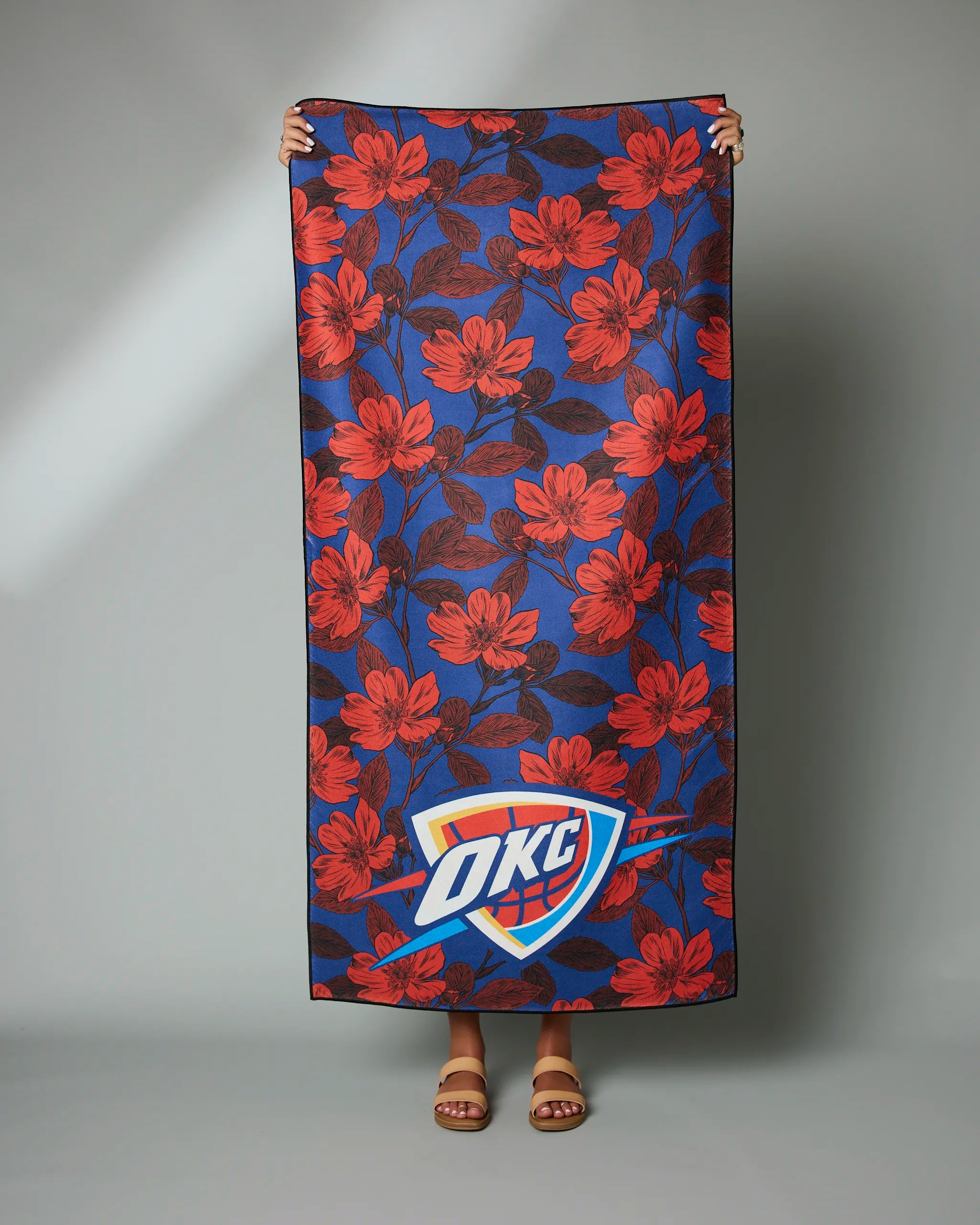 A person holds up the Geometry OKC Floral towel, featuring large red flowers and the OKC Thunder logo; only their hands and sandaled feet are visible.