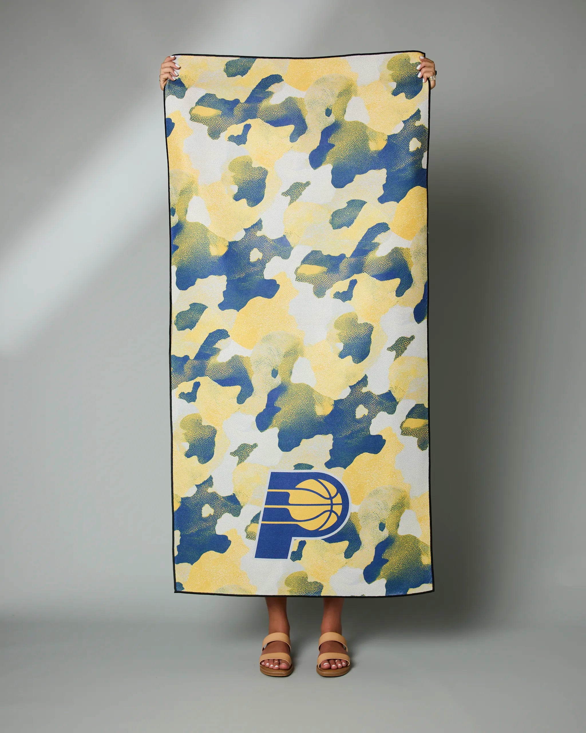 A person holds the Geometry Pacers Camo towel, featuring a blue, yellow, and white camouflage design with an Indiana Pacers logo in one corner. Only their hands and feet are visible.