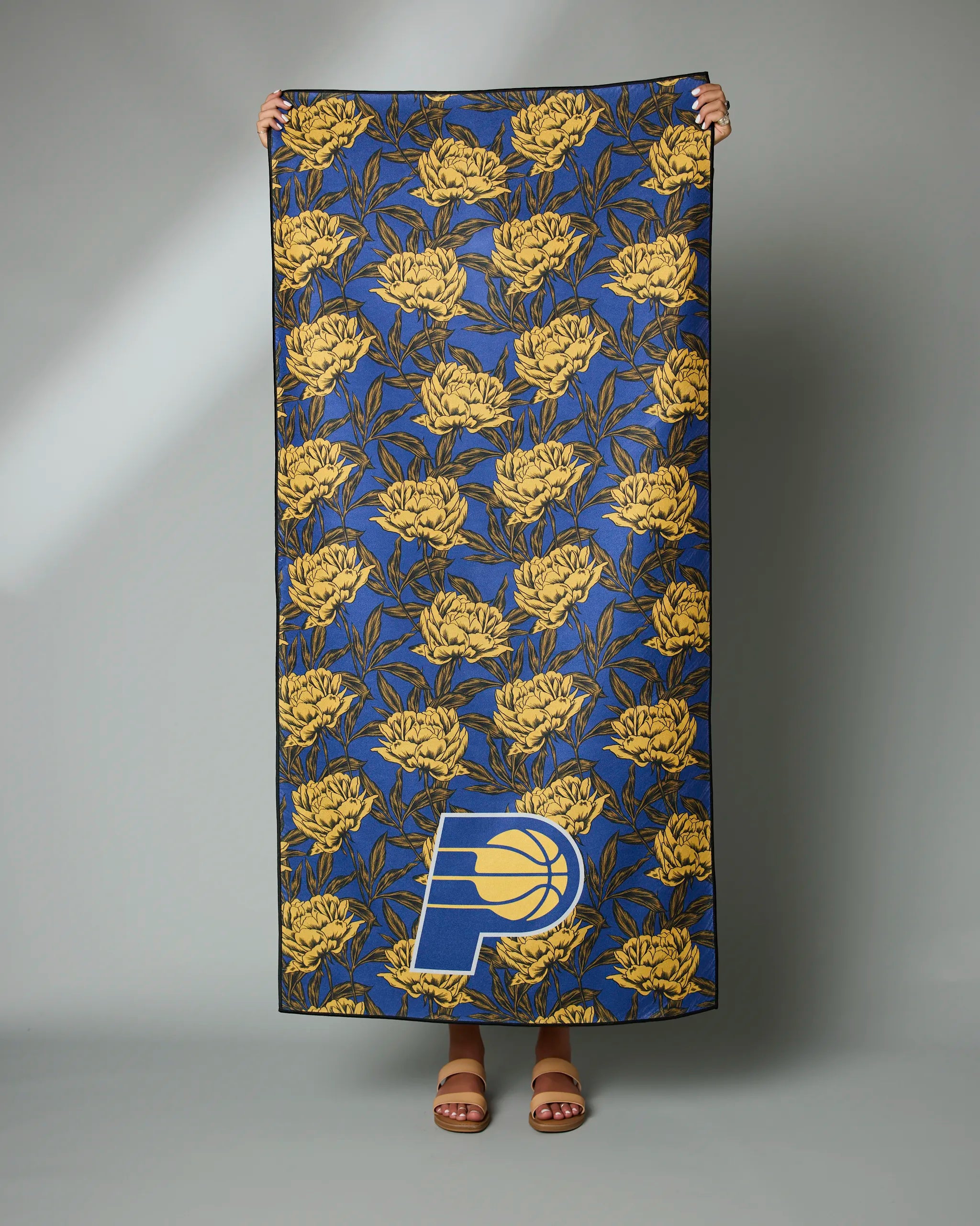 Only the persons hands and feet are visible as they hold up the Pacers Floral towel by Geometry, featuring a blue background, yellow floral pattern, and the Indiana Pacers logo on the bottom right.