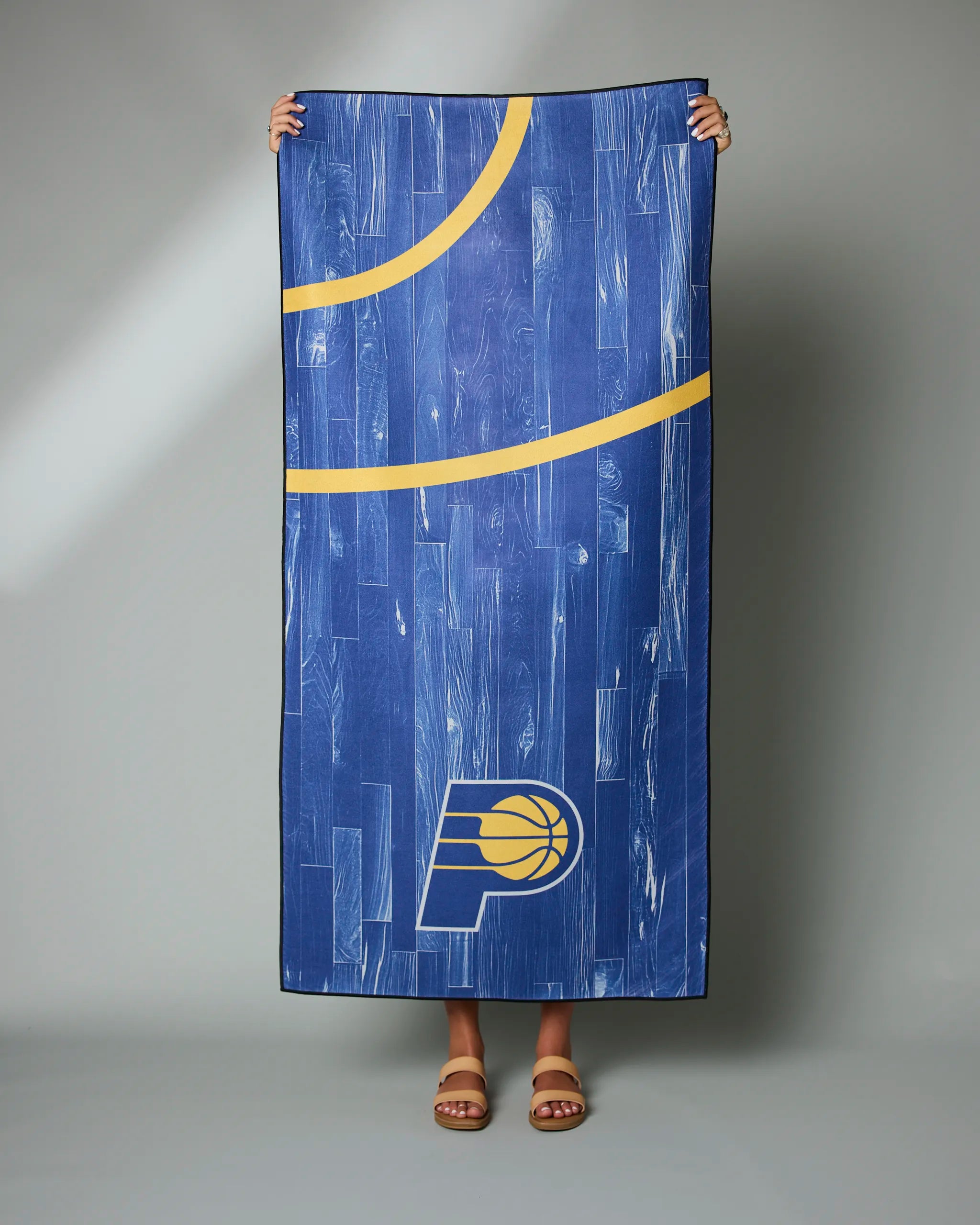 A person displays the blue Pacers Zone towel by Geometry, featuring yellow lines and a basketball logo inspired by the Indiana Pacers, against a plain gray background; only their feet and hands are visible.