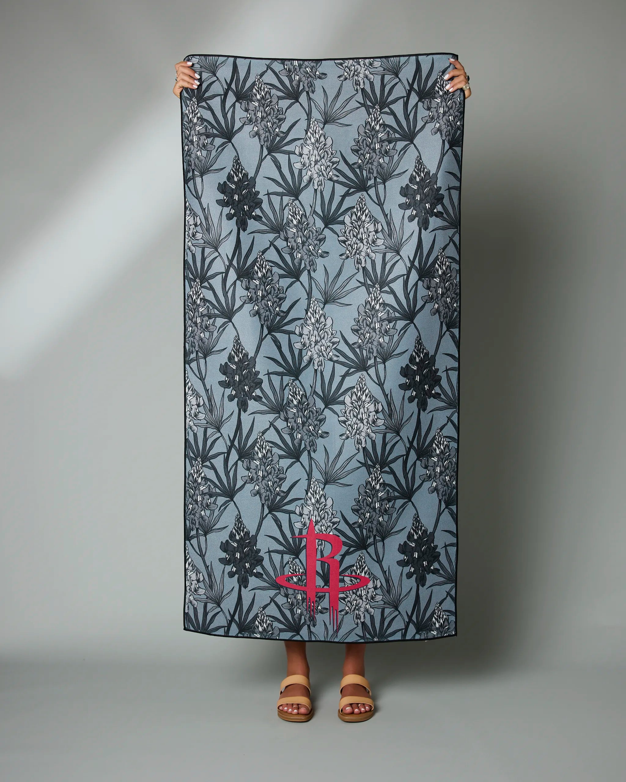 A person holds up the Geometry Rockets Floral towel, featuring a blue-gray background, black and white floral pattern, and a red R logo near the bottom; only their hands and sandaled feet are visible.