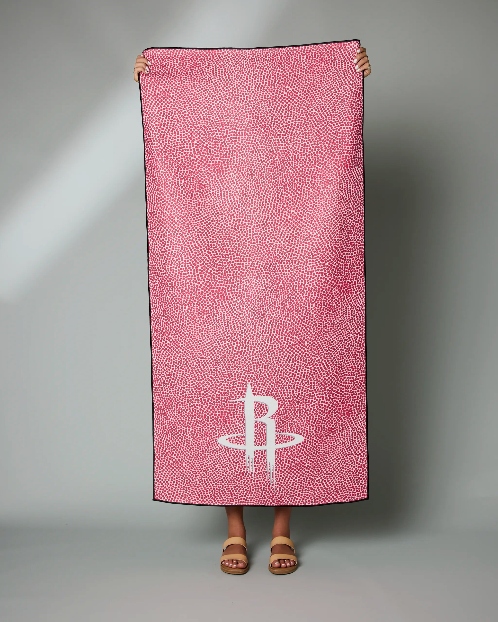 A person stands holding the Geometry Rockets Grip, a large pink towel with small white dots and a white Houston Rockets logo near the bottom, covering their body except for sandal-clad feet. The background is plain gray.