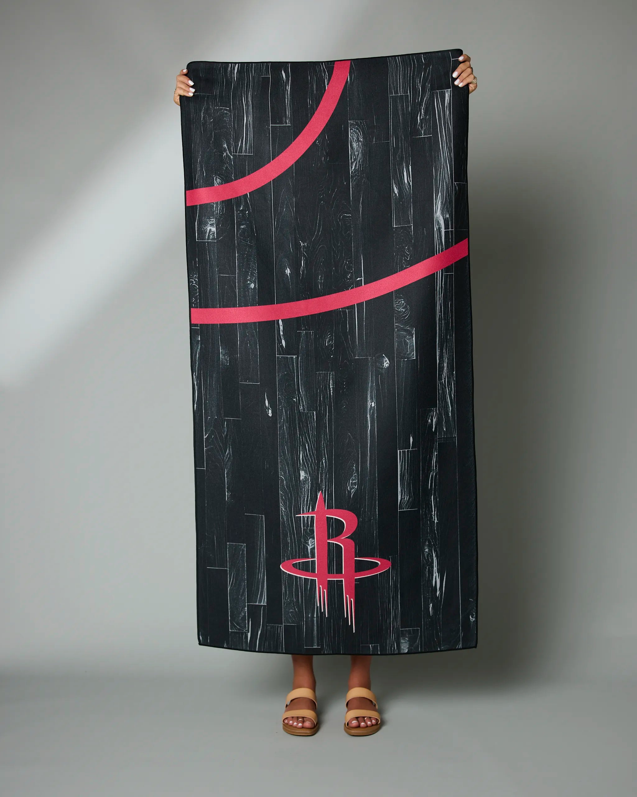 A person holds up the Geometry Rockets Zone towel, featuring red lines and a red Houston Rockets logo on black, against a gray background. Only their hands and feet in brown sandals are visible.