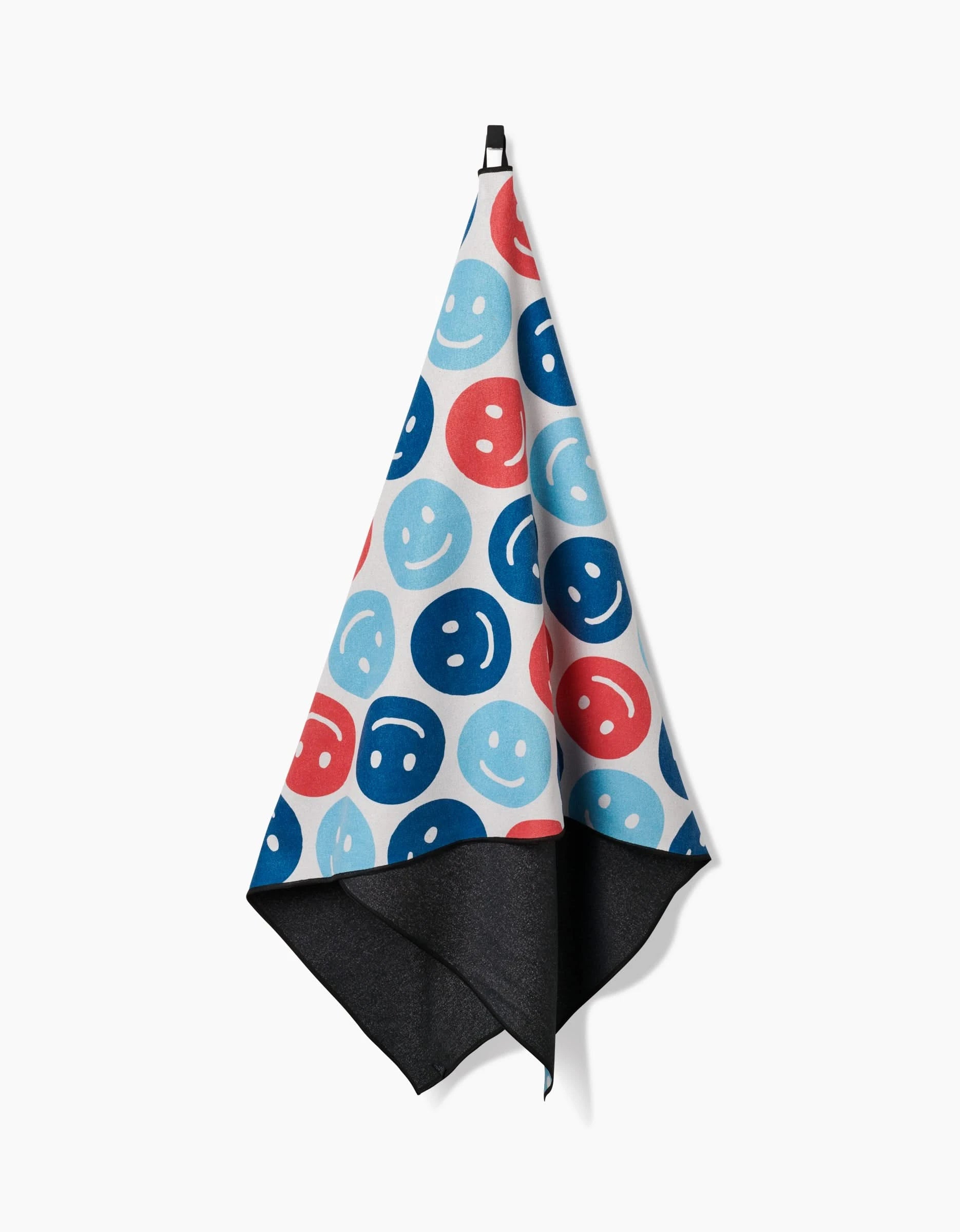 The Hufton Studio Smiley Face July beach towel, featuring blue, red, and light blue smiley faces with a solid black inner side, hangs on a hook against a white background. It is quick-drying.