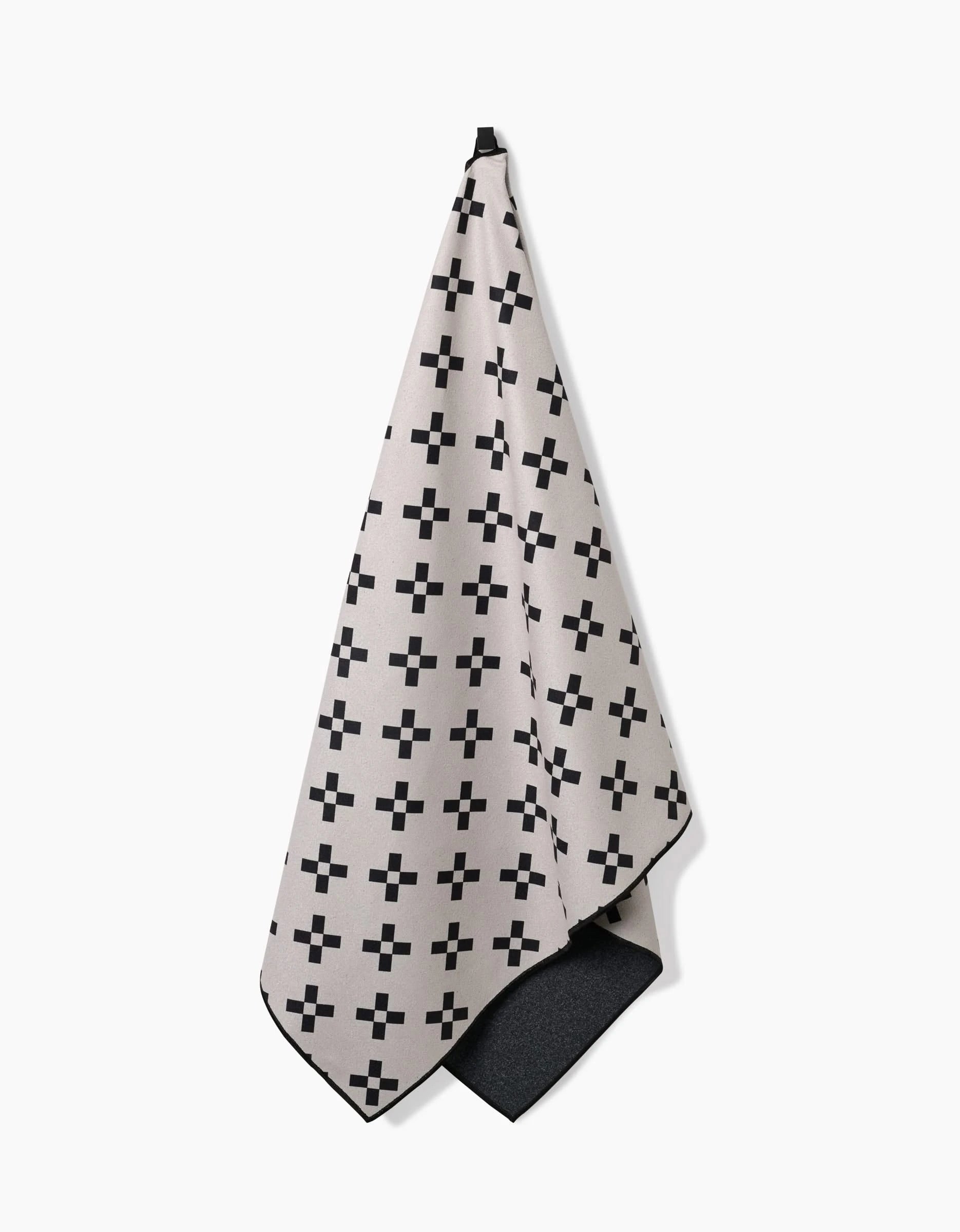 The Tova Beach Towel by Anna Christian, in beige with a black geometric plus sign pattern, hangs from a hook. Crafted from recycled materials, its corner folds up to show the quick-drying black underside.