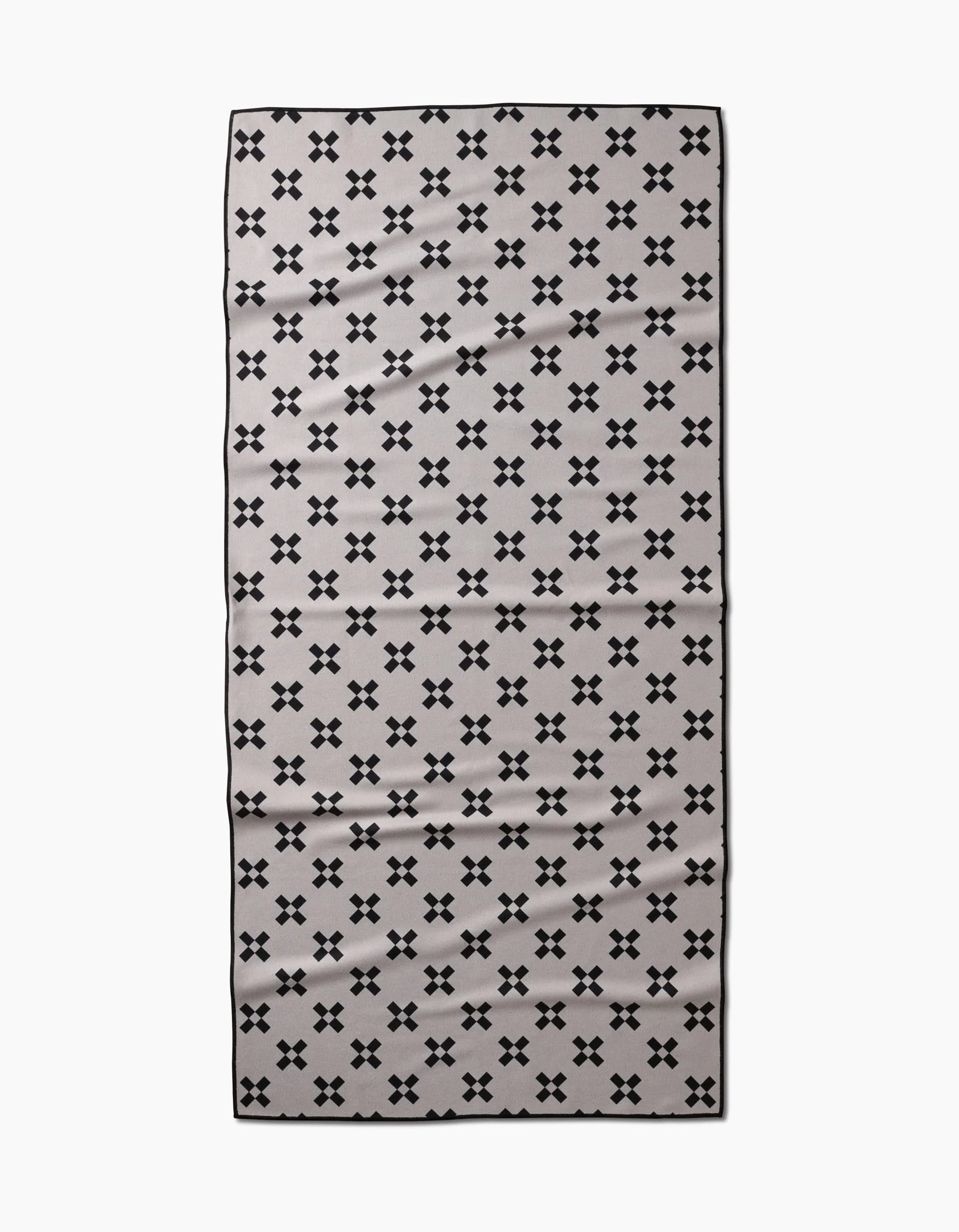 The Tova Beach Towel by Anna Christian is a rectangular, light gray towel with small black X patterns and a thin black border. Made from recycled materials, it is quick-drying and designed for the beach.