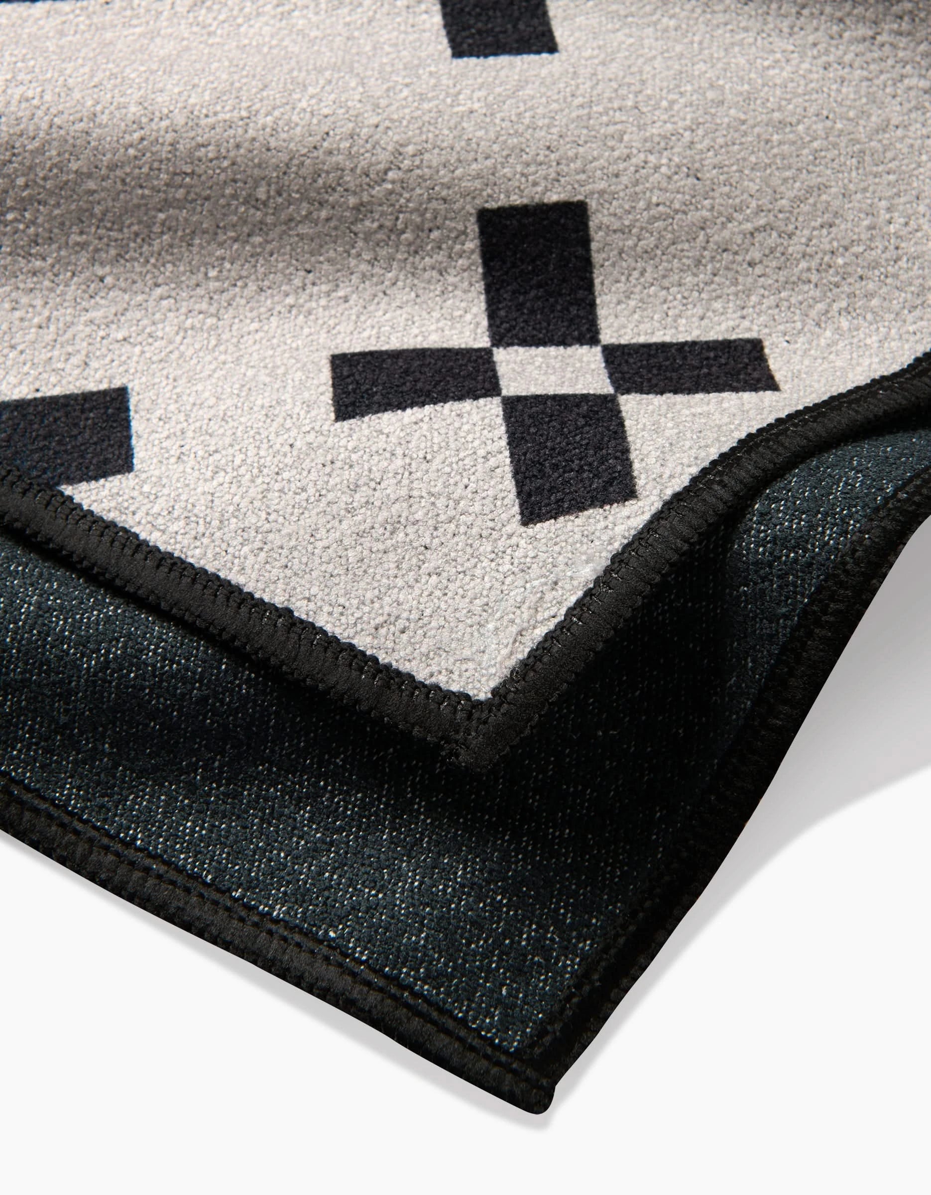 A close-up of the Tova Beach Towel by Anna Christian shows its beige surface with black plus sign patterns, a dark textured reverse, and sleek black trim. Made from recycled materials.