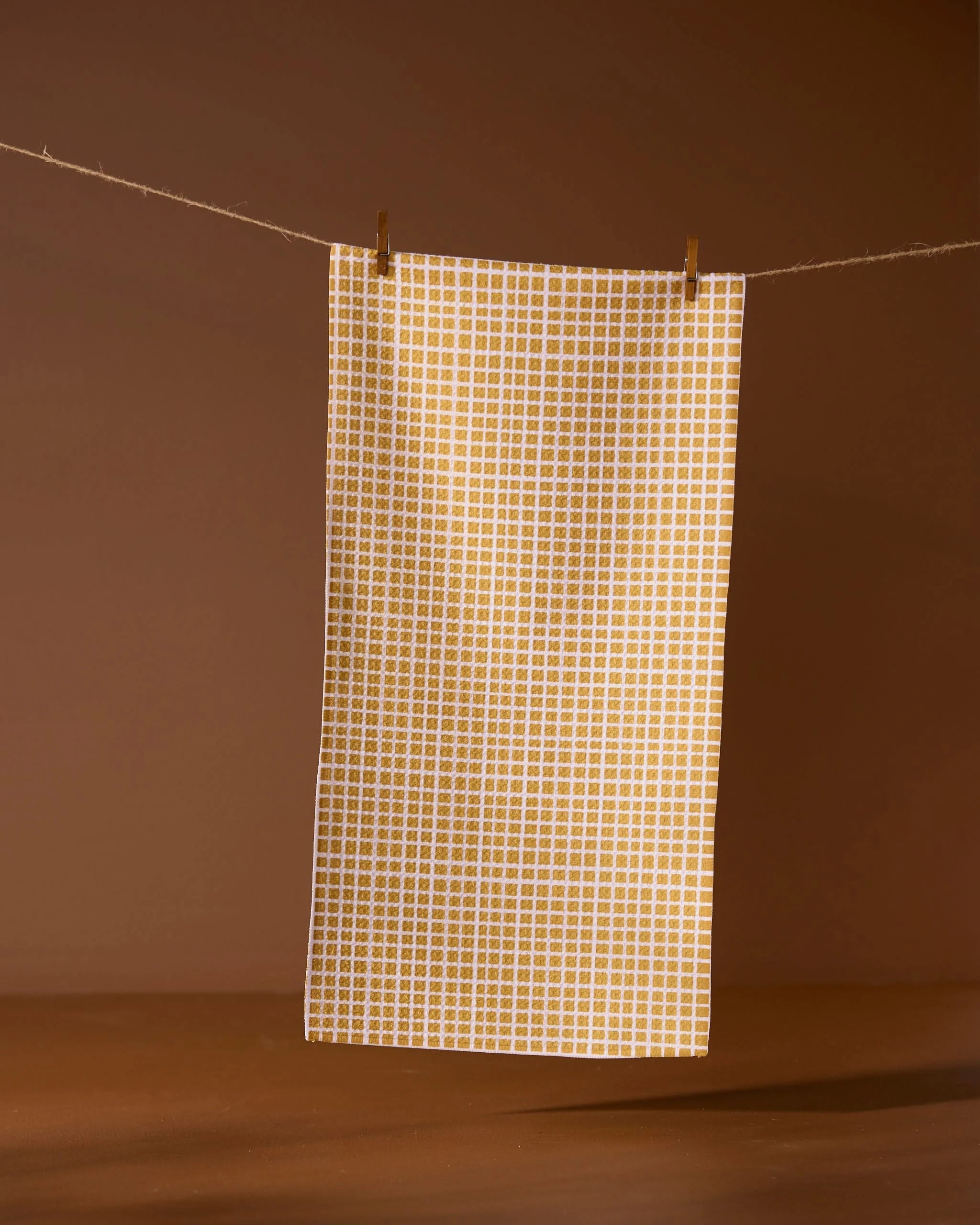 The Geometry Amberline towel, featuring a white and yellow checked design, hangs from a string with two clothespins against a brown background.