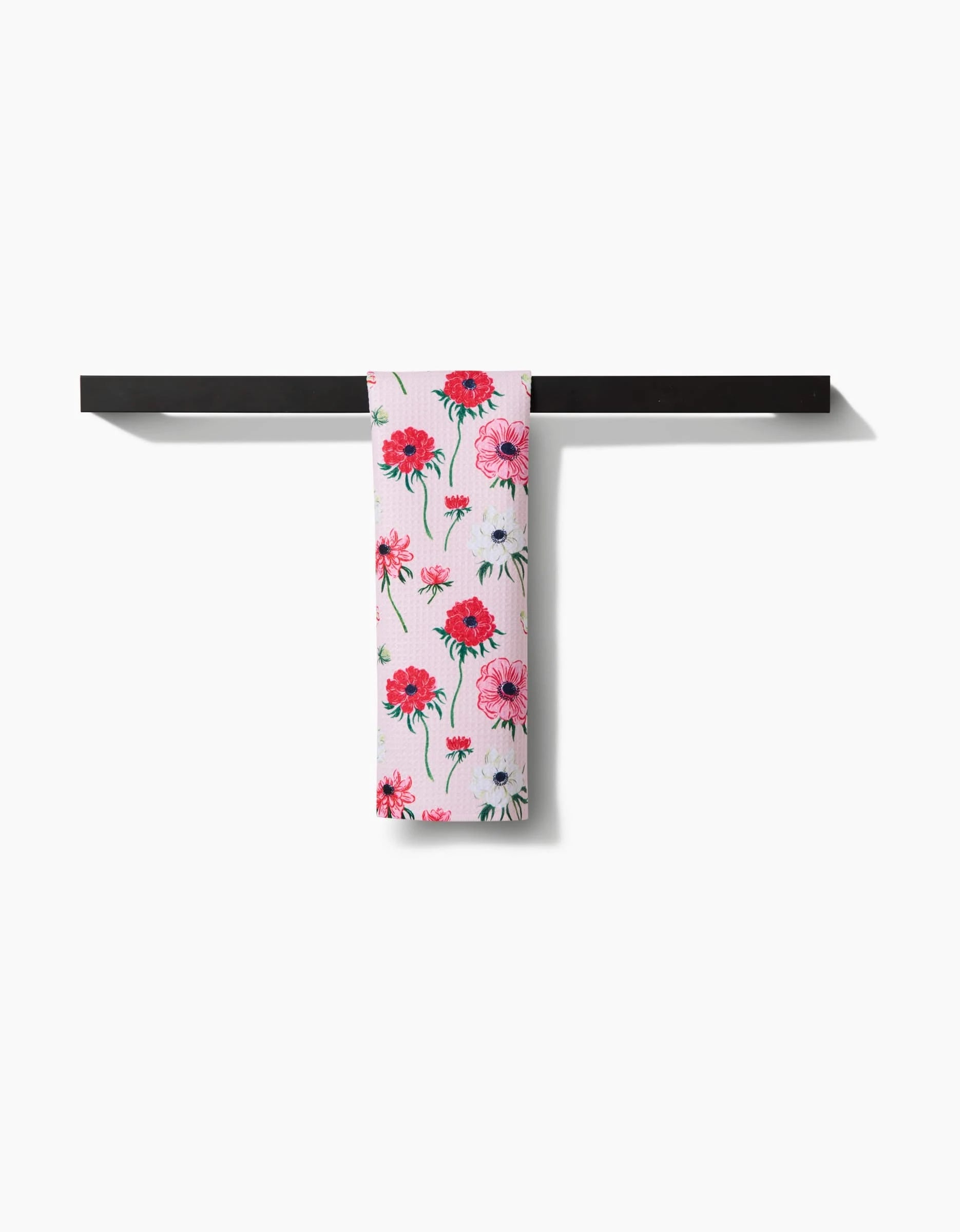 The Linda Fox Anemone Garden Flowers bar towel, featuring a pink and red floral design on white recycled fabric, hangs neatly over a simple black rack against a plain white background.
