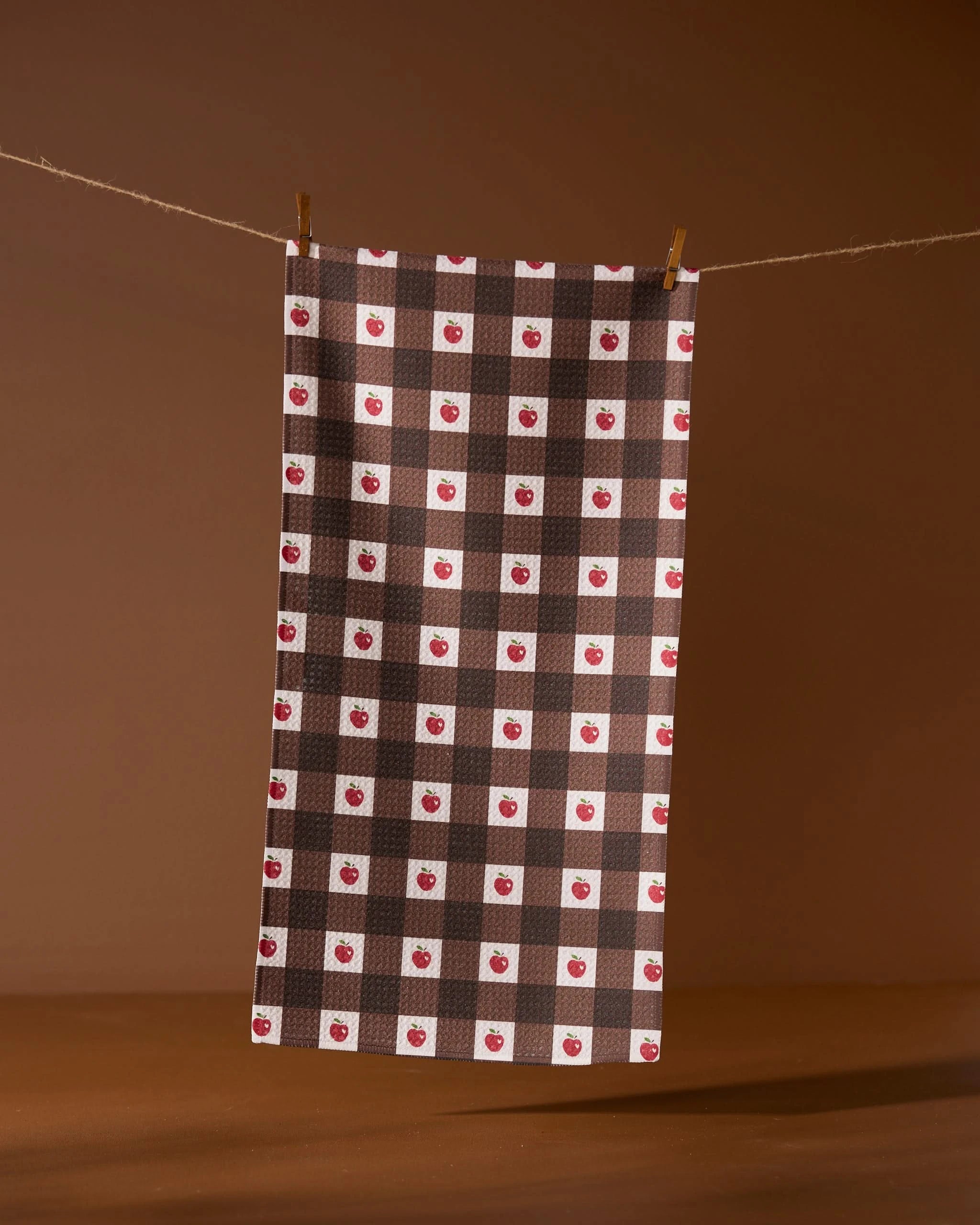 The Geometry Apple Picnic bar towel, ultra-absorbent with red and white checkered squares and small apple accents, hangs from a clothesline against a brown background.