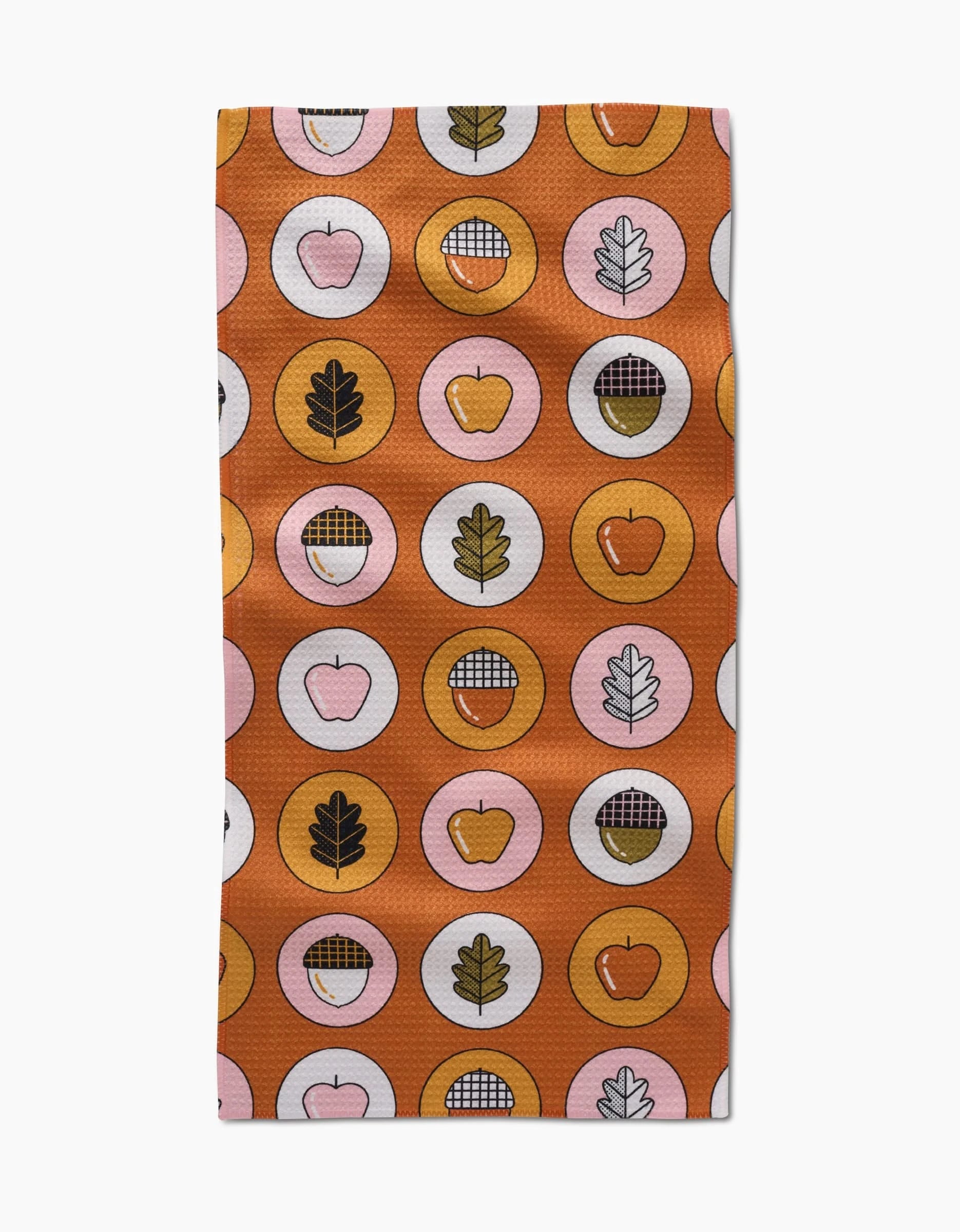 The Apples Acorns recycled kitchen towel by Megan Roy features a pattern of circles on an orange background with apples, pie slices, bowls, and oak leaves in black, white, pink, yellow, and red.