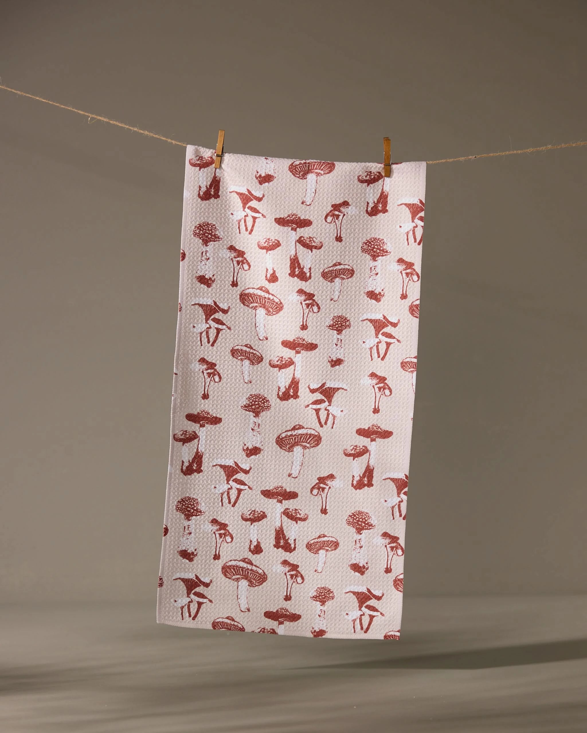 The Geometry Autumn Mushrooms towel, featuring a red mushroom pattern on white, hangs from a clothesline with two wooden clothespins against a neutral background.