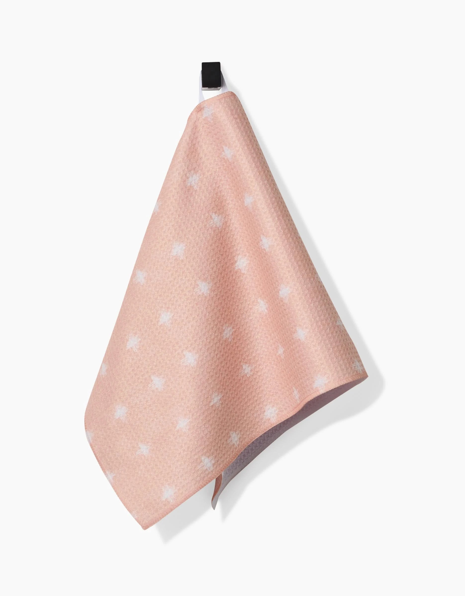 The Amy MacCready Aveline bar towel features a light pink hue with small white star patterns, a subtle waffle texture, and is made from ultra-absorbent Geoweave fabric. It hangs from a black hook against a white background.