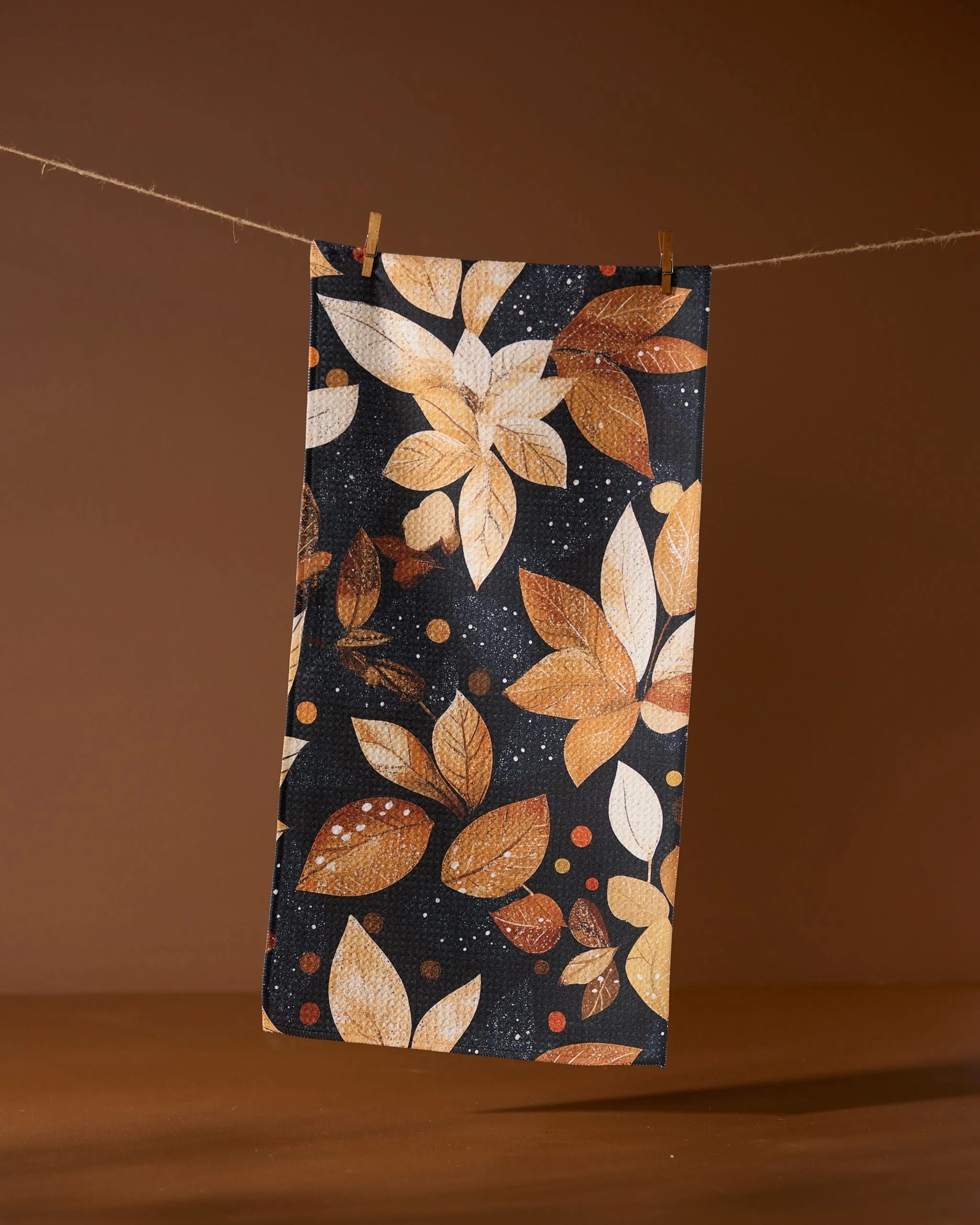 The Geometry Botanical Wonderland bar towel features an autumn leaf design in brown, orange, and cream. Made from recycled materials, it’s eco-friendly and stylish, shown hanging with clothespins against a brown background.
