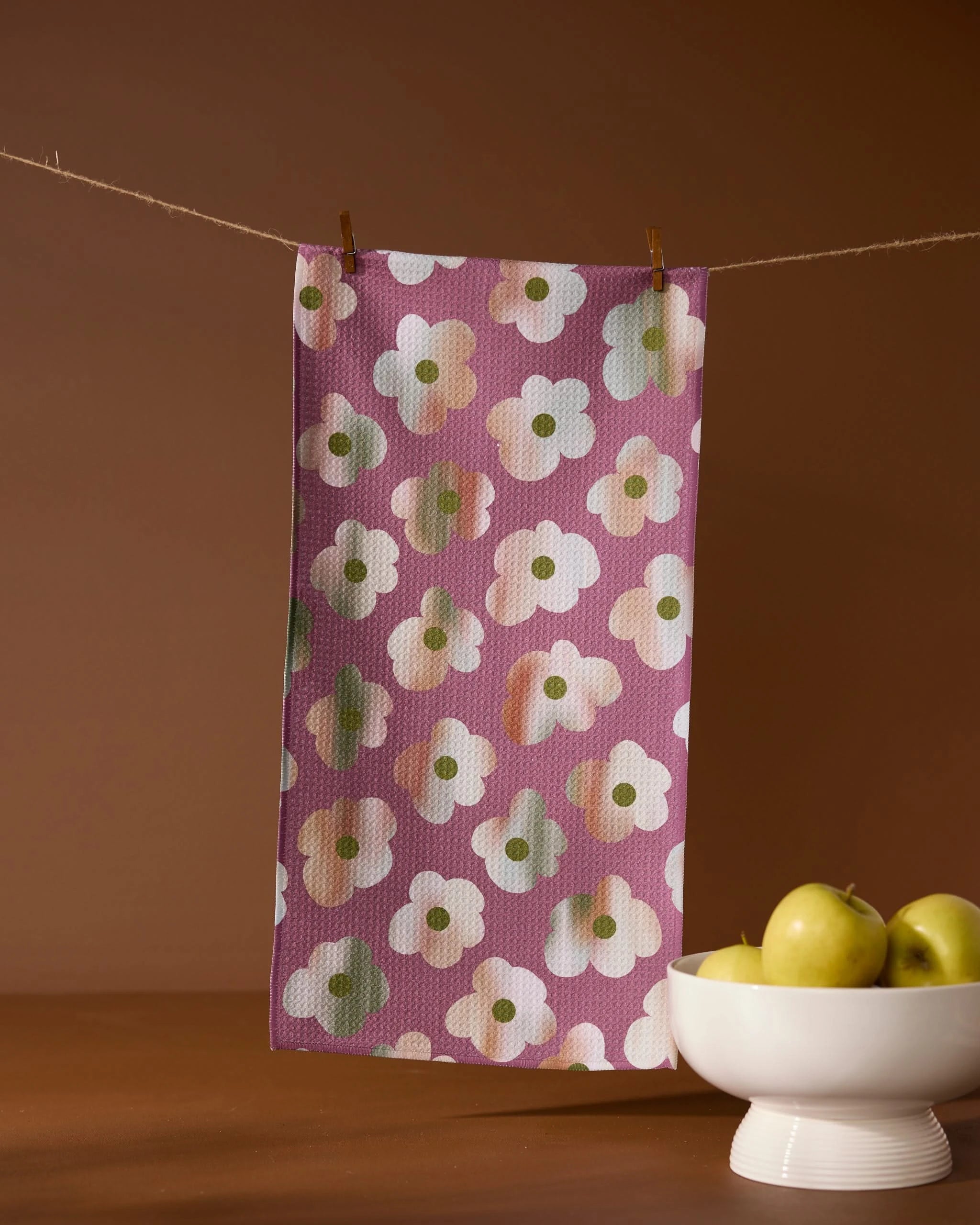 The Brushed Bouquet bar towel by Geometry, featuring a white and yellow floral pattern on recycled purple fabric, hangs on a clothesline beside a white bowl of green apples set on a brown surface with a matching background.