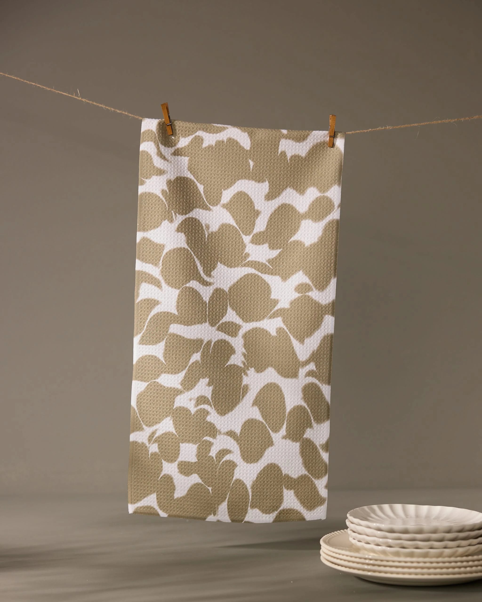 The Geometry Canopy bar towel, featuring a beige and white pattern and ultra-absorbent design, hangs from a string with clothespins. In the foreground, white ceramic plates are stacked neatly on a gray surface.