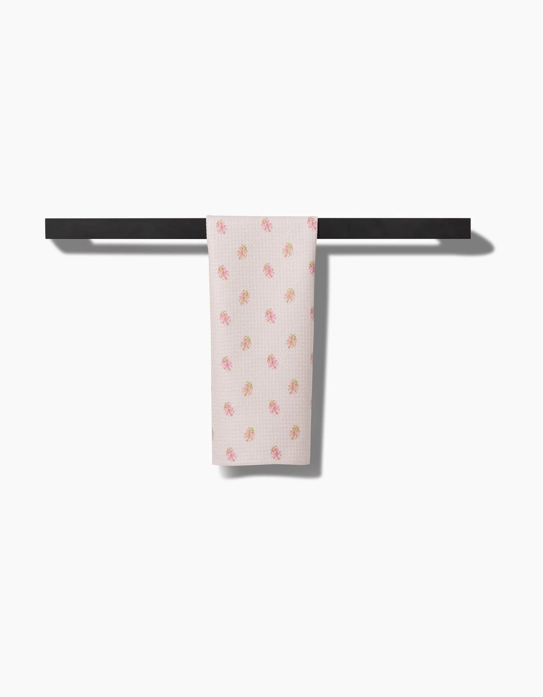 The Cindy bar towel by Kelsi Fullmer, made from recycled materials and decorated with small pink and yellow flowers, hangs neatly on a simple black towel bar against a plain white background.