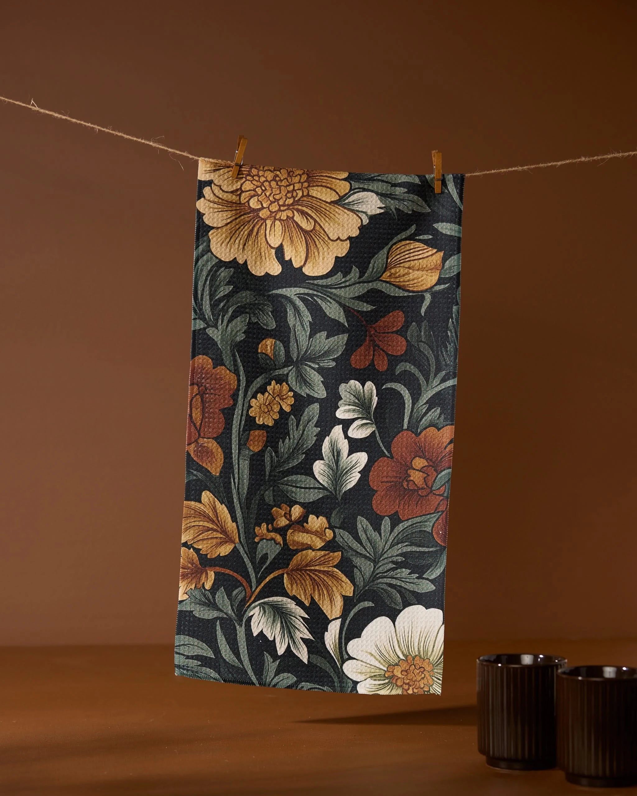The Geometry Classic Foliage bar towel, featuring yellow, orange, and white florals on a dark background and made from recycled materials, hangs on a clothesline with two dark cups placed below to the right.