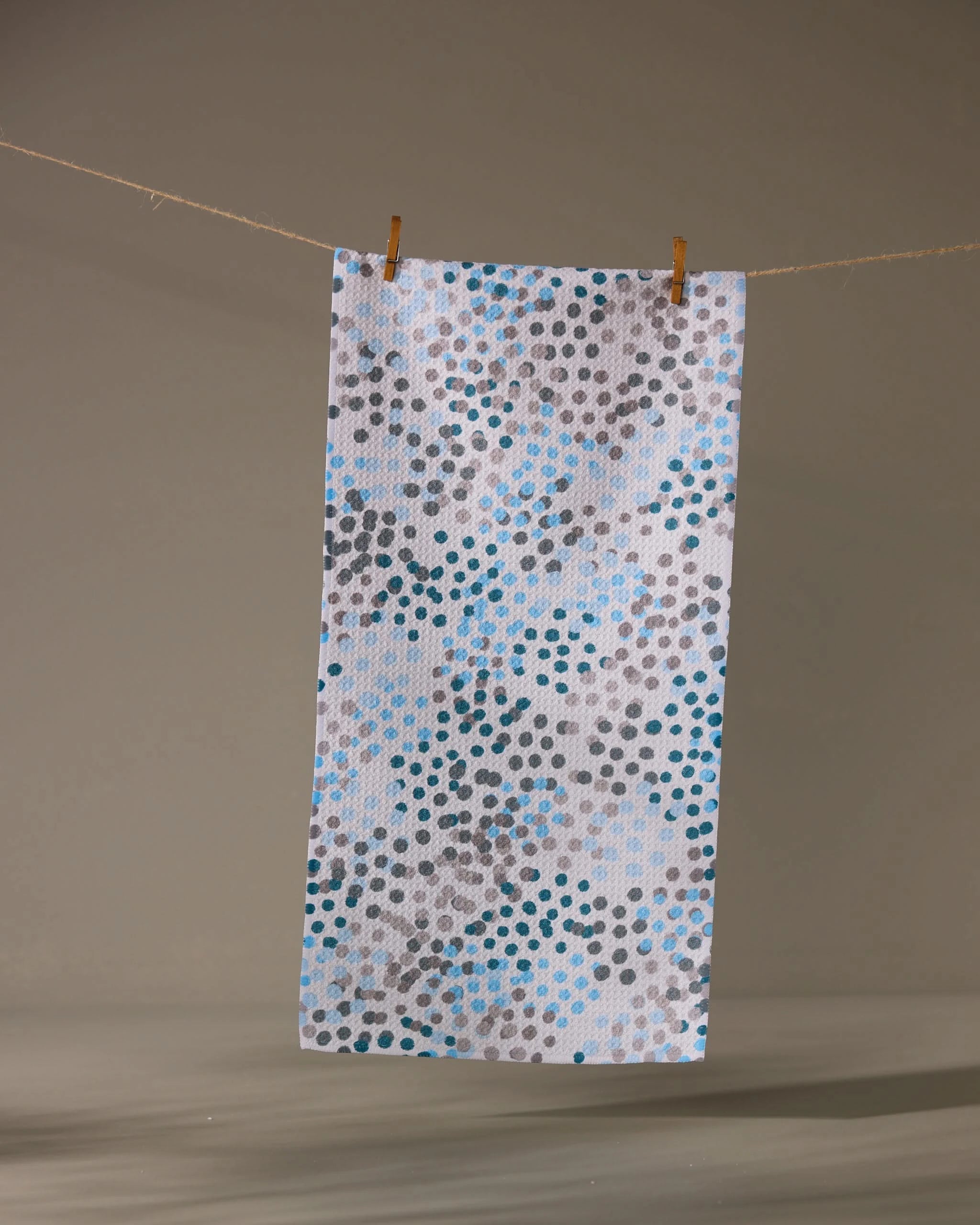 The Geometry Cool Dots recycled kitchen towel, white and rectangular with blue, grey, and black dot patterns, hangs from a string by two wooden clothespins against a simple background.