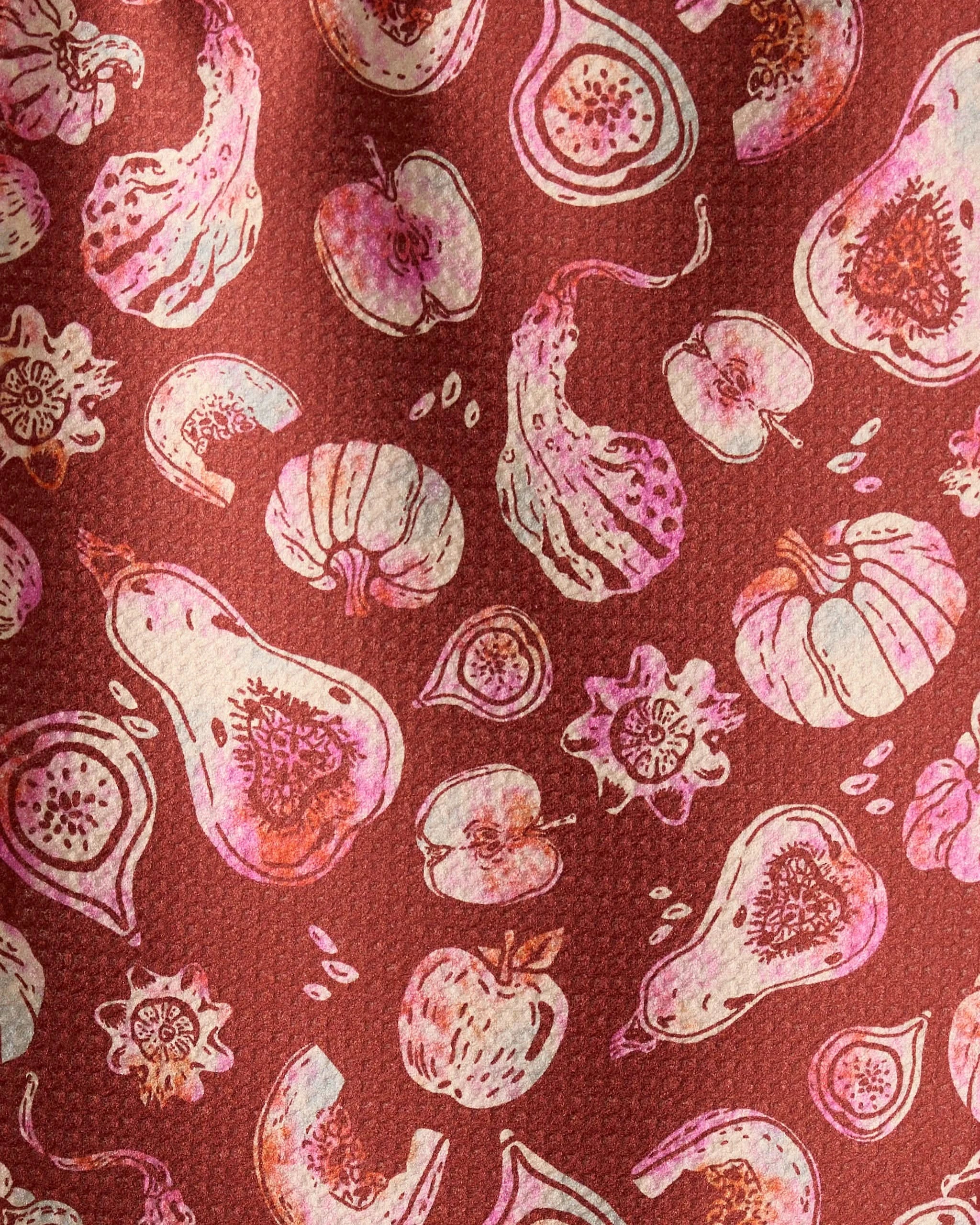 Geometry’s Cosecha bar towel features illustrations of fruits and vegetables—like gourds, figs, pumpkins, and apples—in pink and cream on a textured reddish-brown background. Made from ultra-absorbent recycled materials.