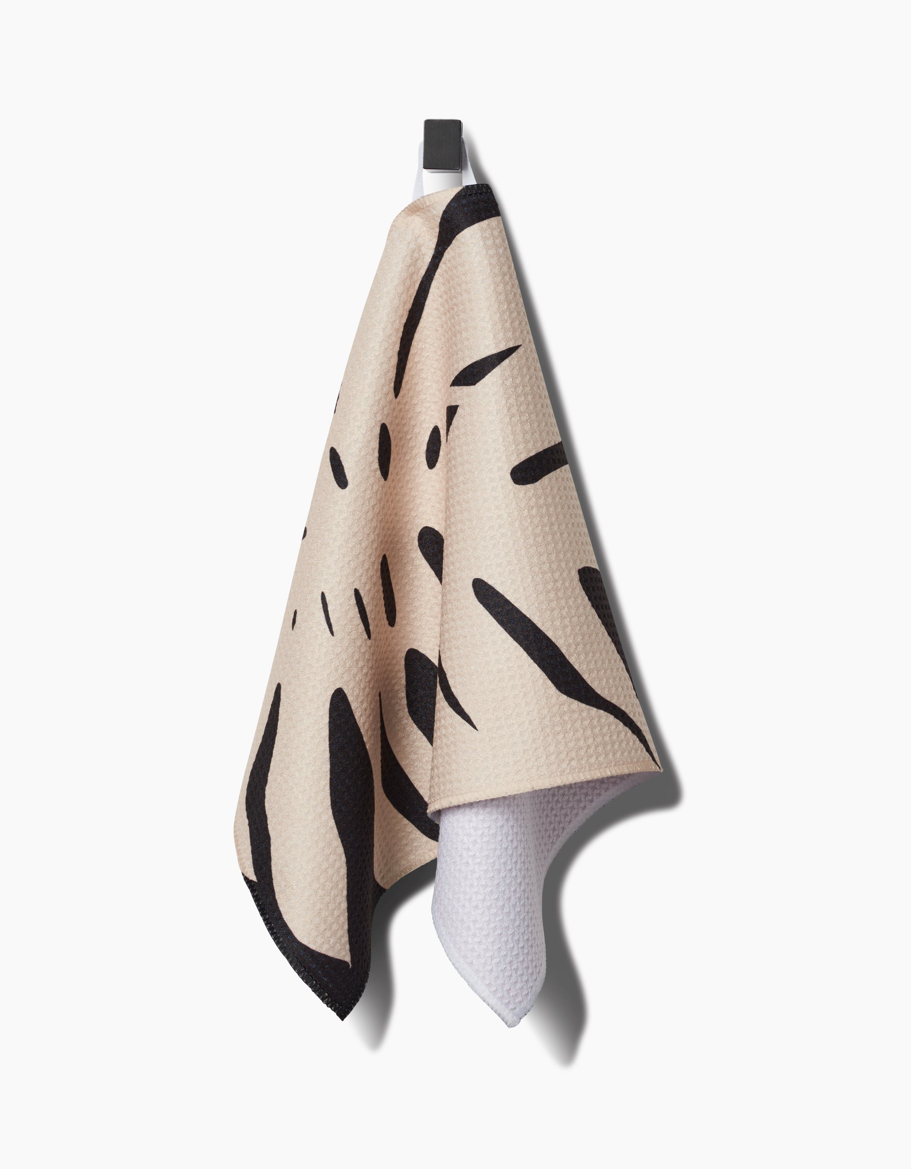 A Cream Monstera bar towel by PrintedPlant with black abstract patterns hangs from a dark hook on a white background. Its waffle-like texture comes from Geoweave fabric, and the reverse side is white.
