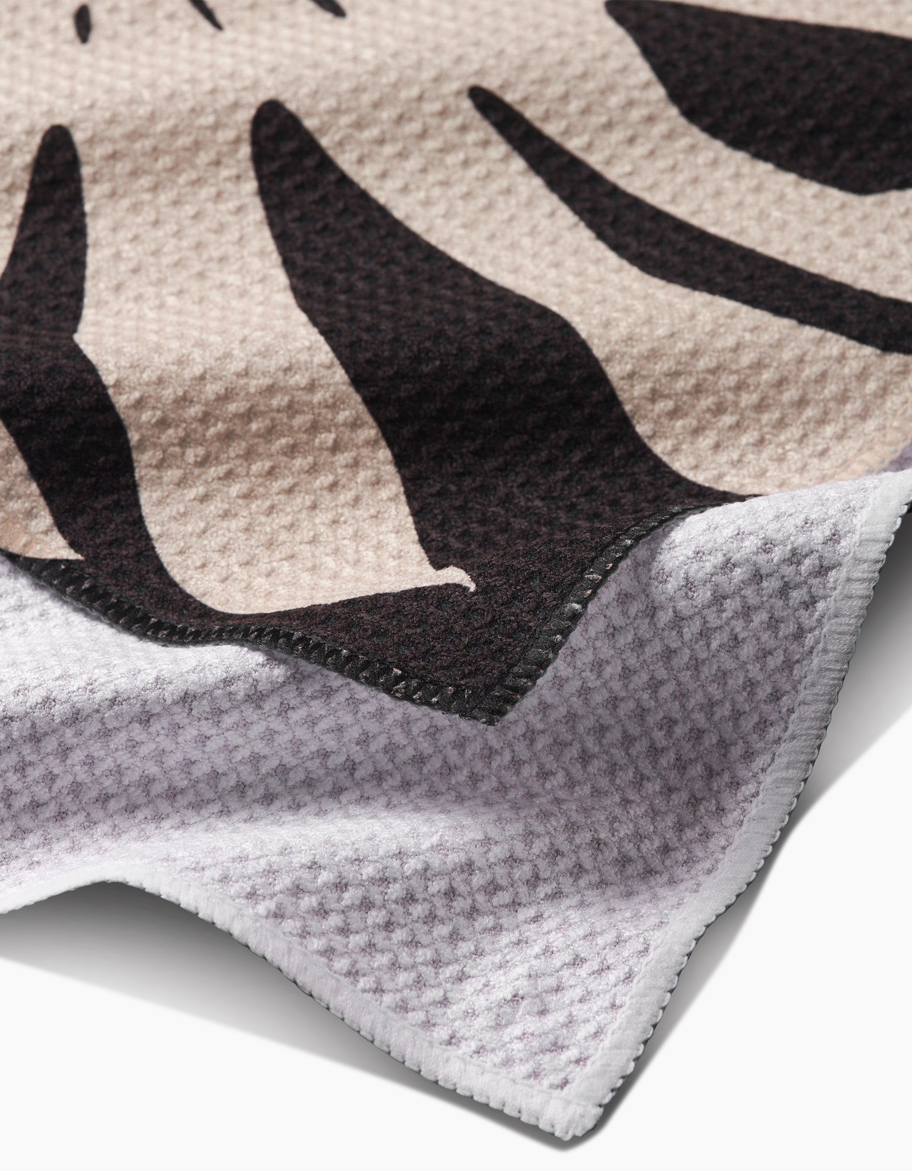 Close-up of the PrintedPlant Cream Monstera Bar Towel made with Geoweave fabric, showcasing a beige and black abstract design on top, a white waffle-like underside, and detailed stitching along the edge.