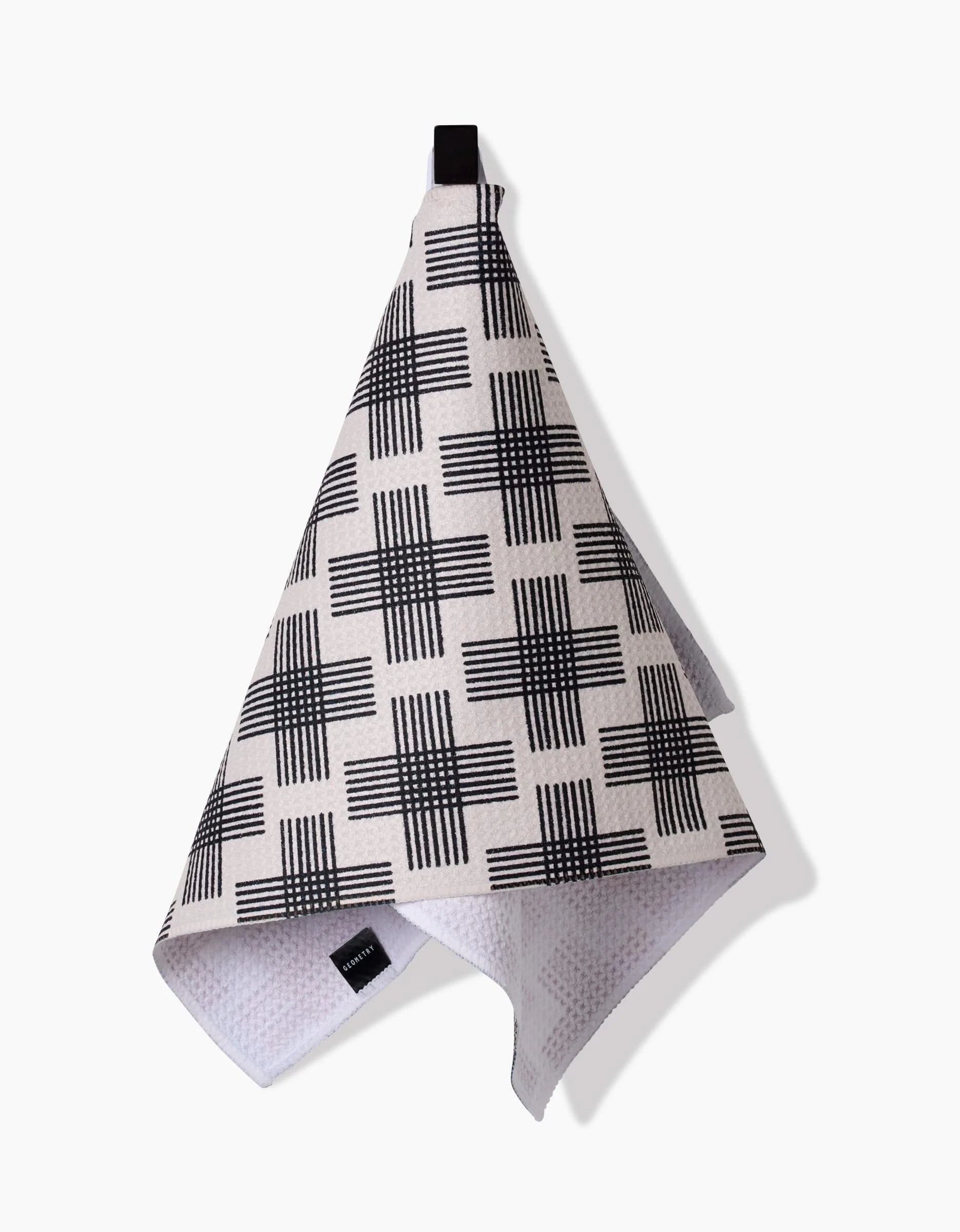 The Rosa Kichline Cross Hatch bar towel in beige features a black geometric check pattern, a black hanging loop, and is crafted from Geoweave fabric, shown against a white background.