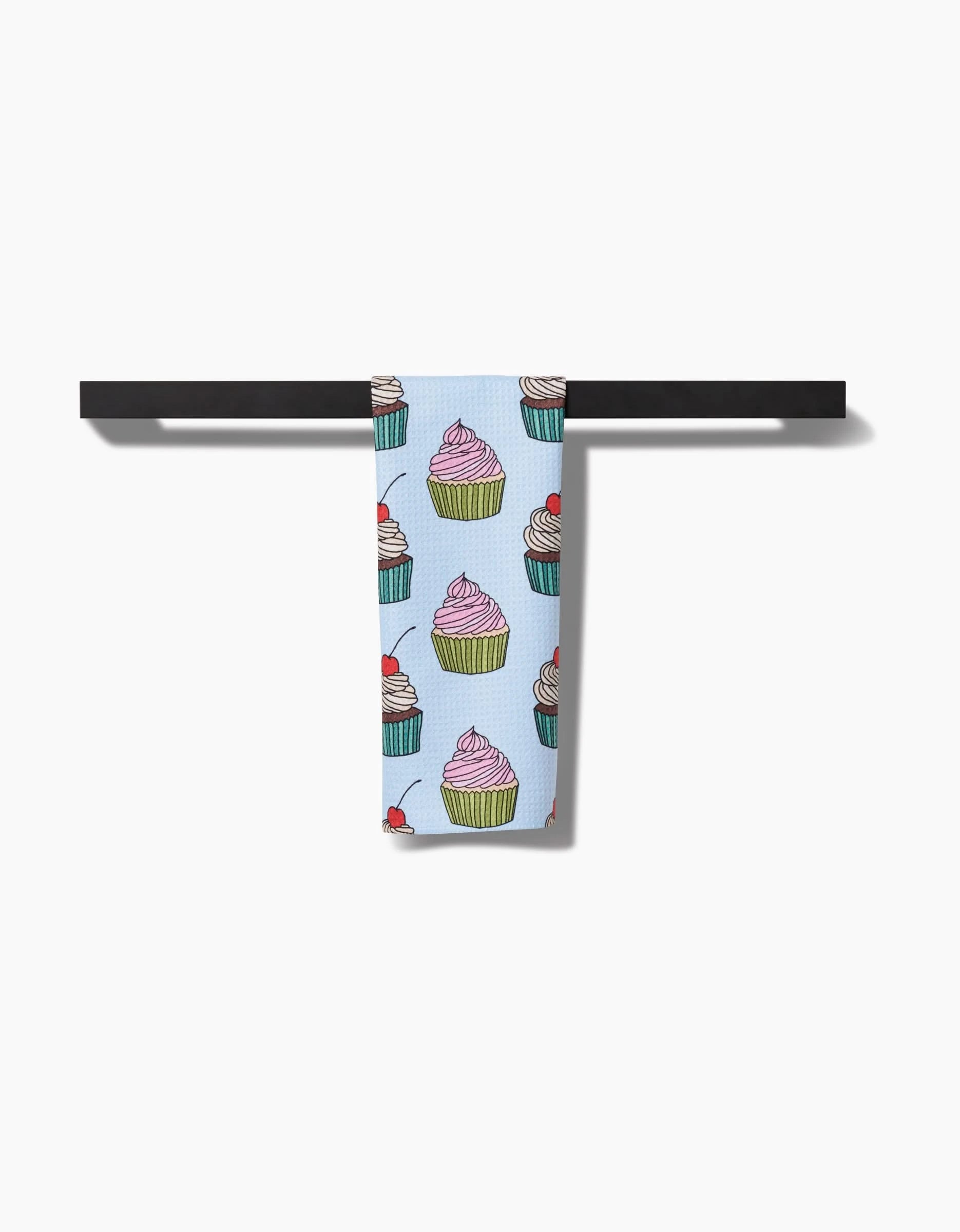The Cupcake Love Bar Towel by Amy Droubay, crafted from Geoweave fabric and adorned with vibrant cupcake designs, is draped neatly over a horizontal black bar set against a white background.