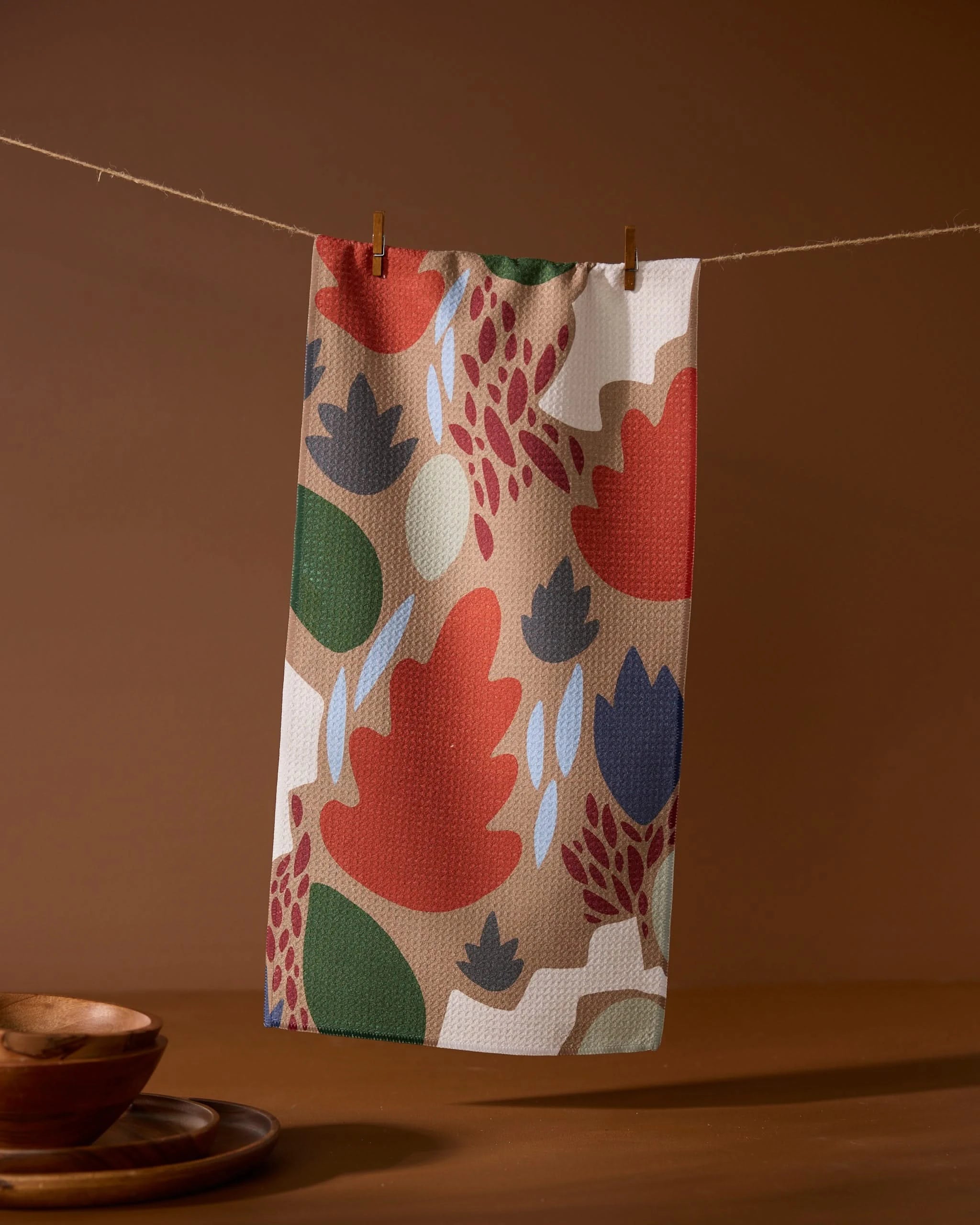 The Geometry Cut Paper Leaves Mocha towel, featuring bold leaf and abstract patterns in red, blue, green, and beige, hangs on a clothesline with wooden clothespins above brown bowls set on a brown surface.