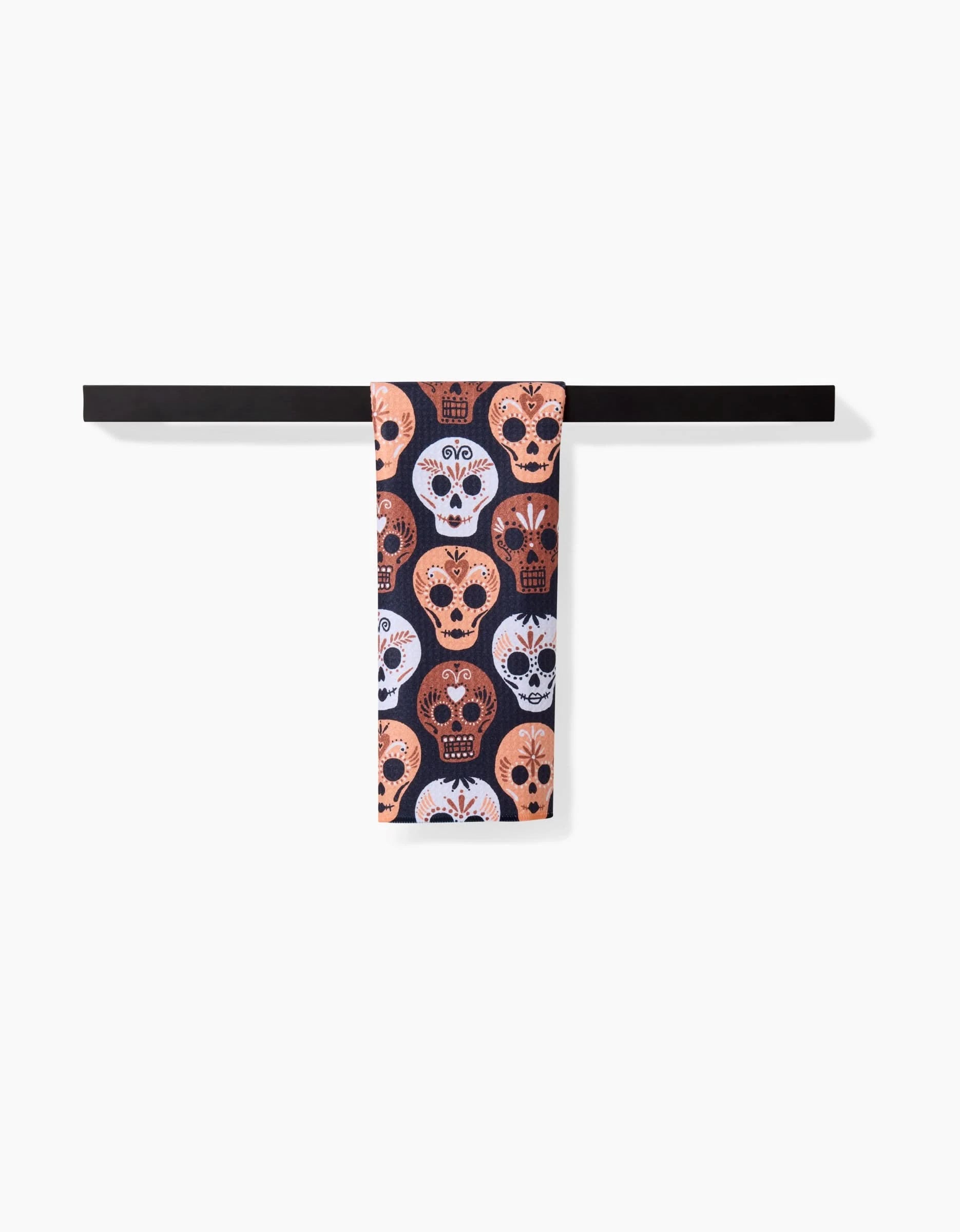 The Day Of The Dead Skulls bar towel by Hufton Studio, featuring vibrant pink, white, and blue sugar skulls, hangs on a black towel bar against a plain white background.