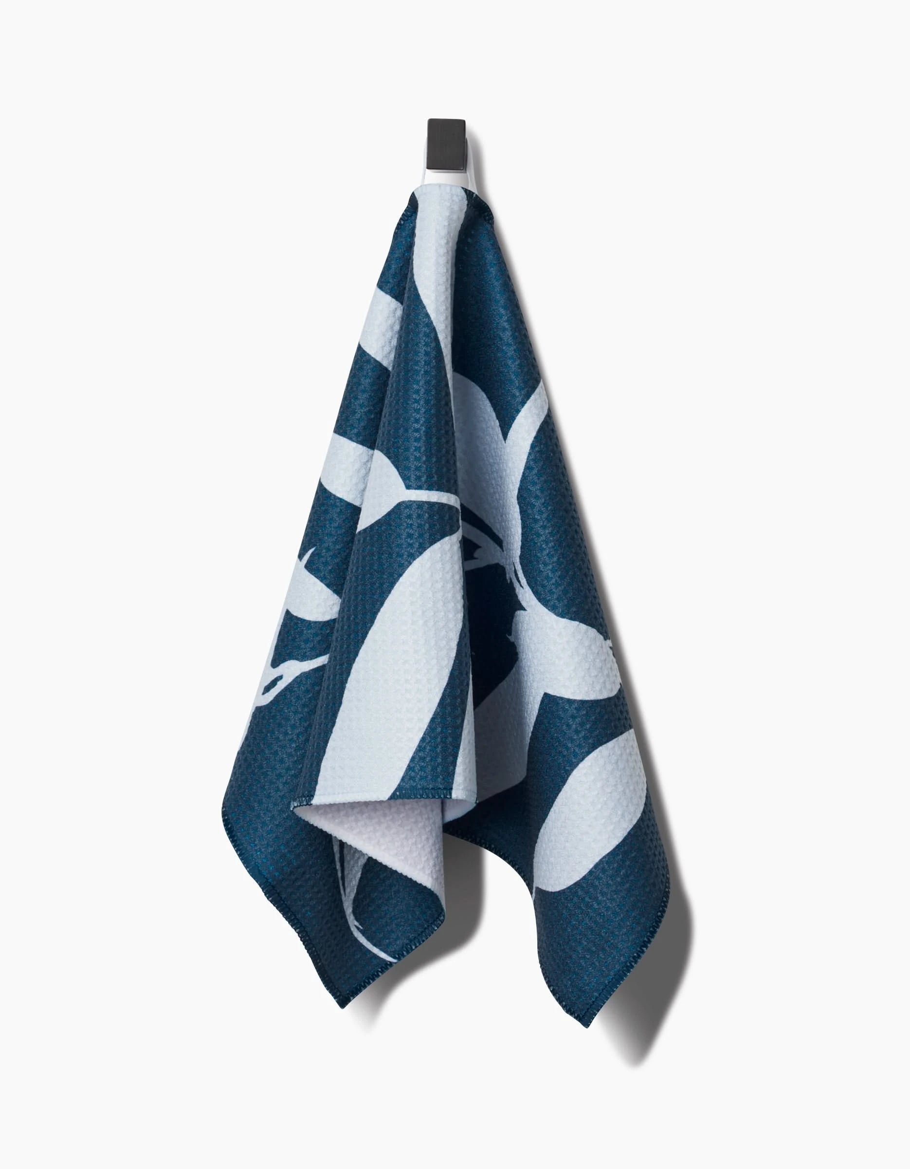 The Doghobble towel by PrintedPlant, made from recycled materials with a bold blue and white leaf pattern, hangs from a black hook on a plain white background.