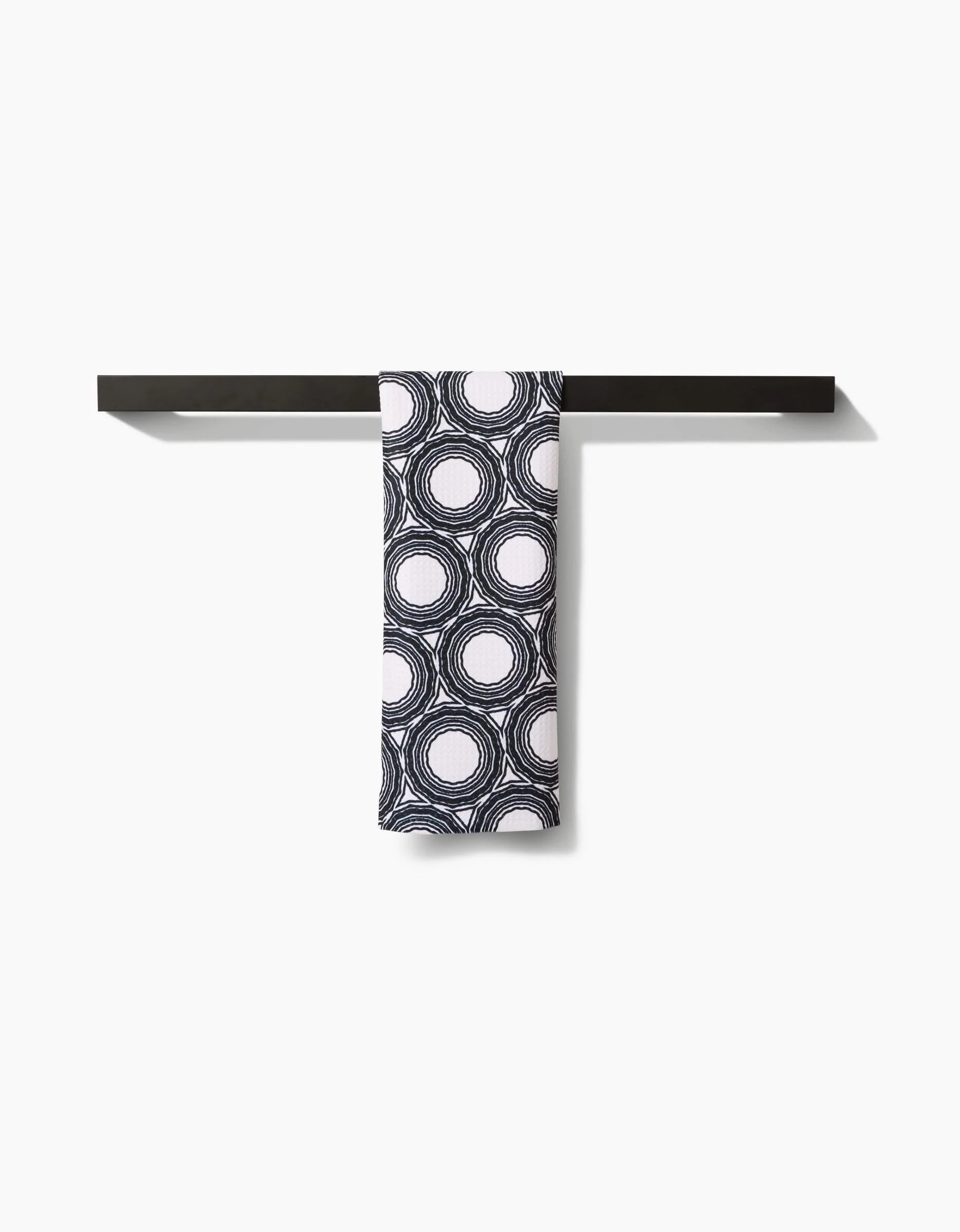 The Dosido Bar Towel by Katherine Berkeley features a black and white circular pattern, crafted from premium Geoweave fabric, and hangs neatly on a minimalist black bar against a plain white background.
