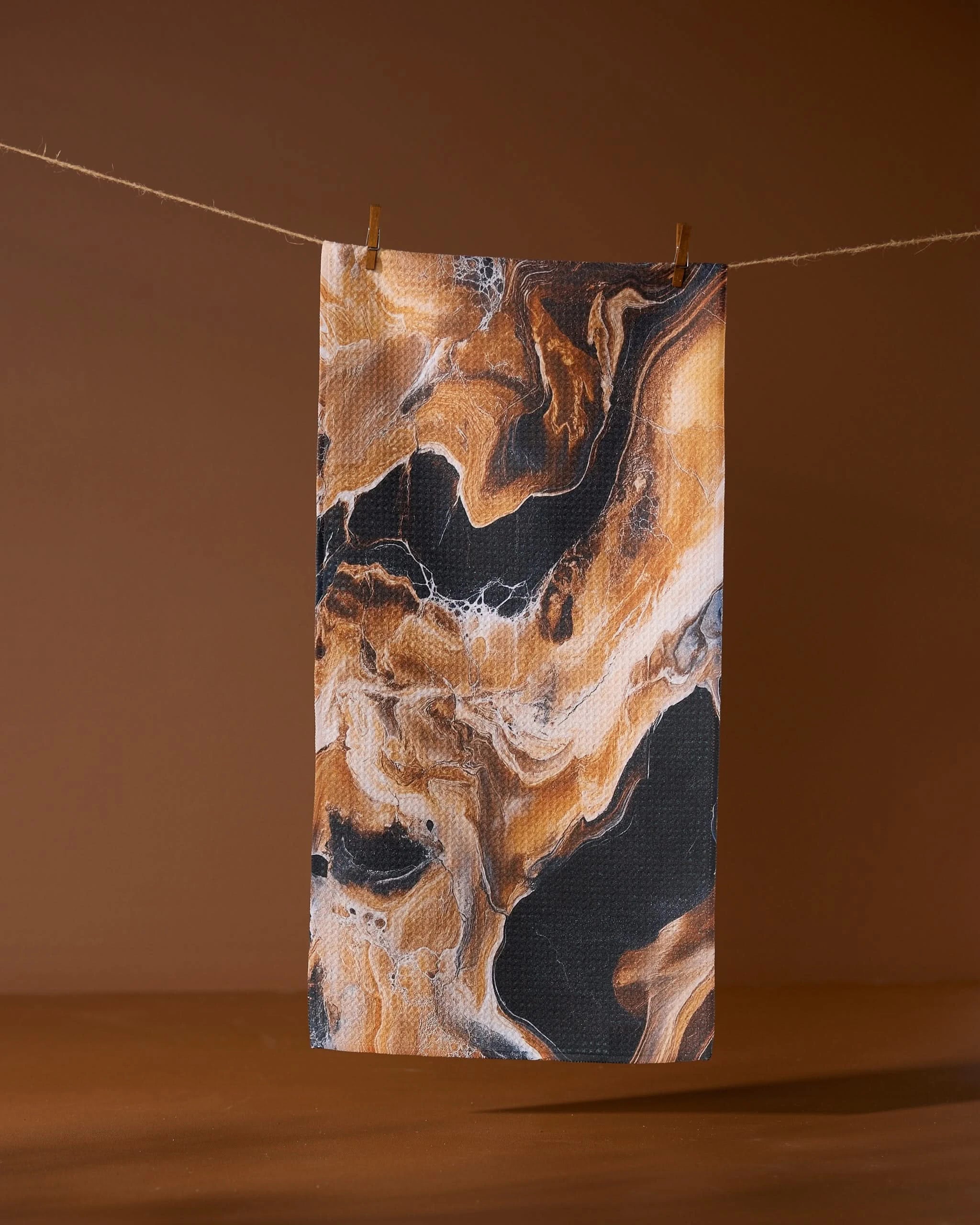 The Geometry Emperador rectangular recycled kitchen towel, featuring a black, brown, and beige marbled pattern, hangs from a string with two clothespins against a solid brown background.