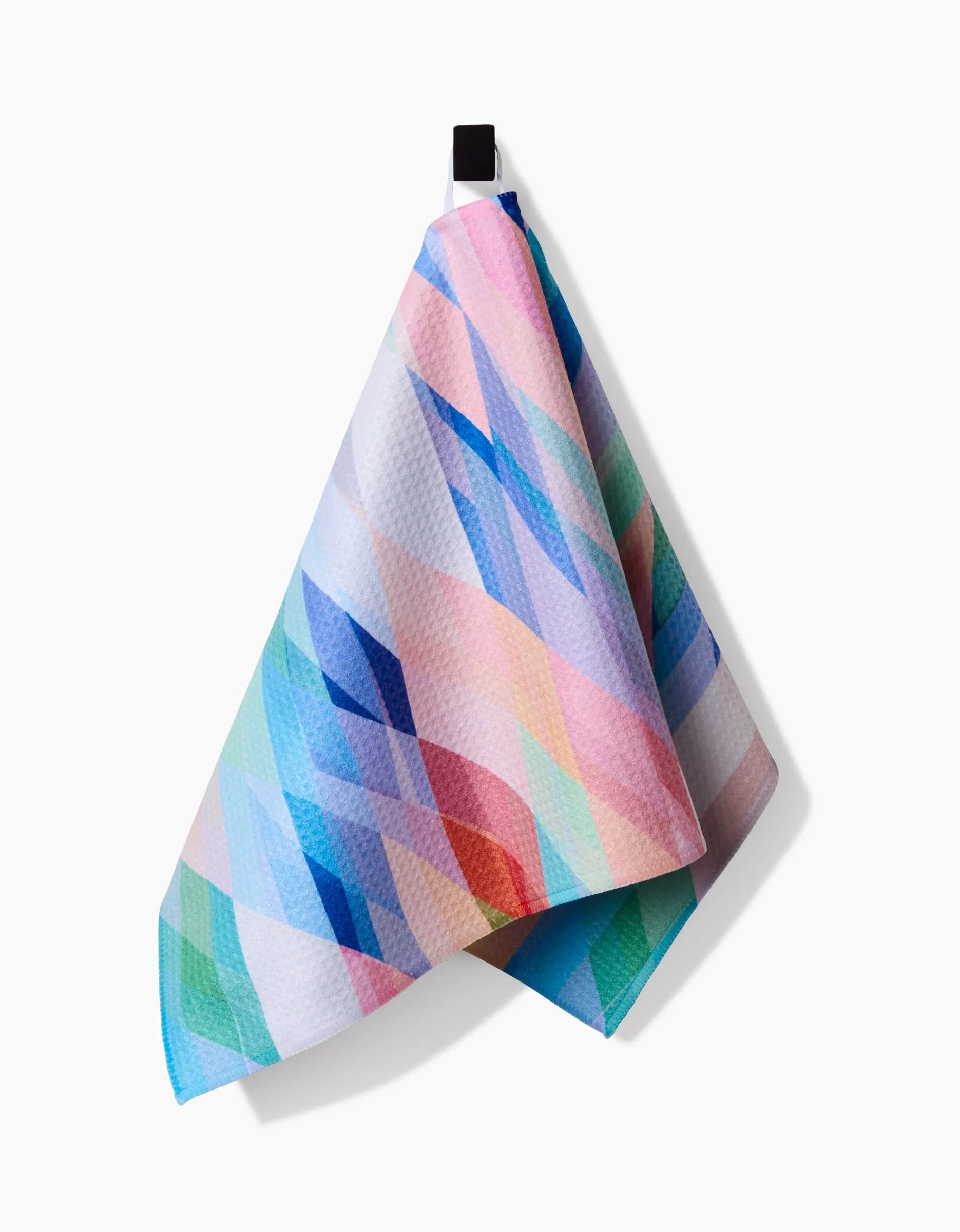The Eventide Bar Towel by Rachel Parker features a geometric diamond pattern in pastel blue, pink, green, and purple, made from recycled materials, and hangs from a black loop against a white background.