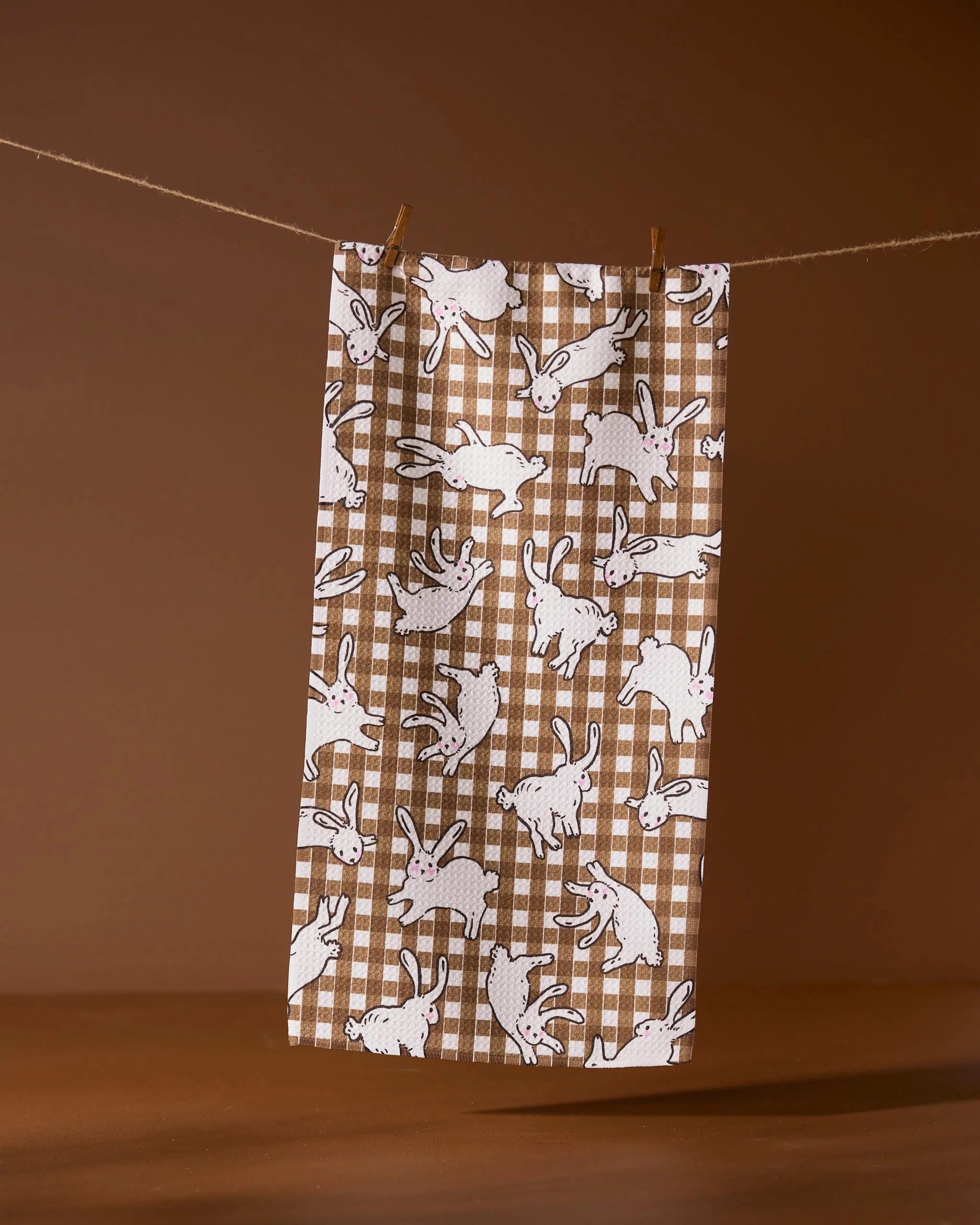 The Fall Bunny Hops recycled kitchen towel by Geometry features a brown and white gingham pattern with playful white rabbits, hanging on a clothesline against a brown background.