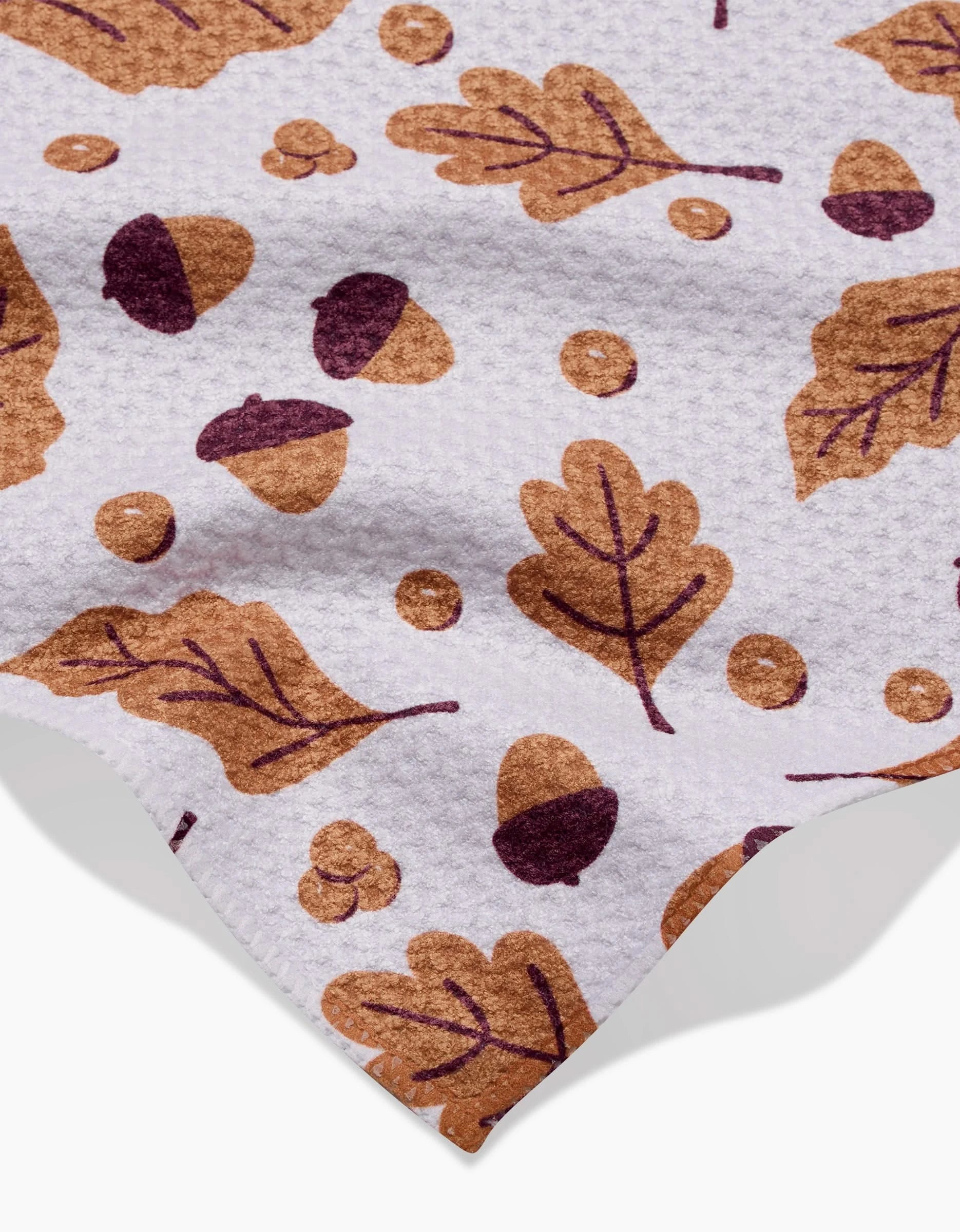 Falling For Fall by Charlie Rowan Designs is a white textured recycled kitchen towel featuring brown and dark purple acorns, oak leaves, and berries, with part of the cloth edge visible.
