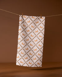 The Geometry Foxes and Twinkles tea towel, crafted from recycled materials, displays a repeating pattern of orange foxes and brown geometric shapes, shown hanging on a clothesline with a brown background.
