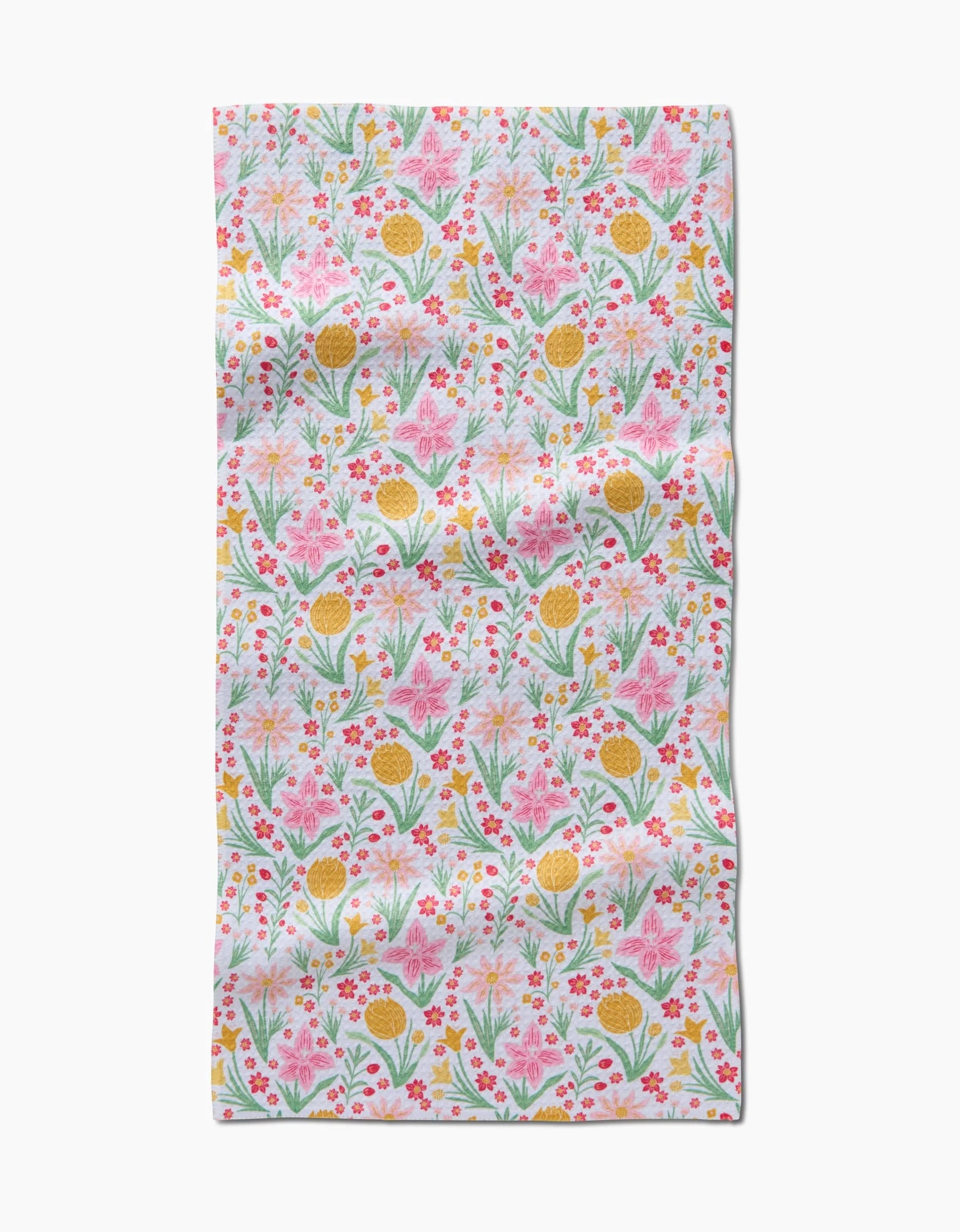 The Garden of Light bar towel by Danelys Sidron features a colorful floral pattern—pink, yellow, and orange flowers with green leaves—on white Geoweave fabric made from recycled materials. The material has a slightly wrinkled look.