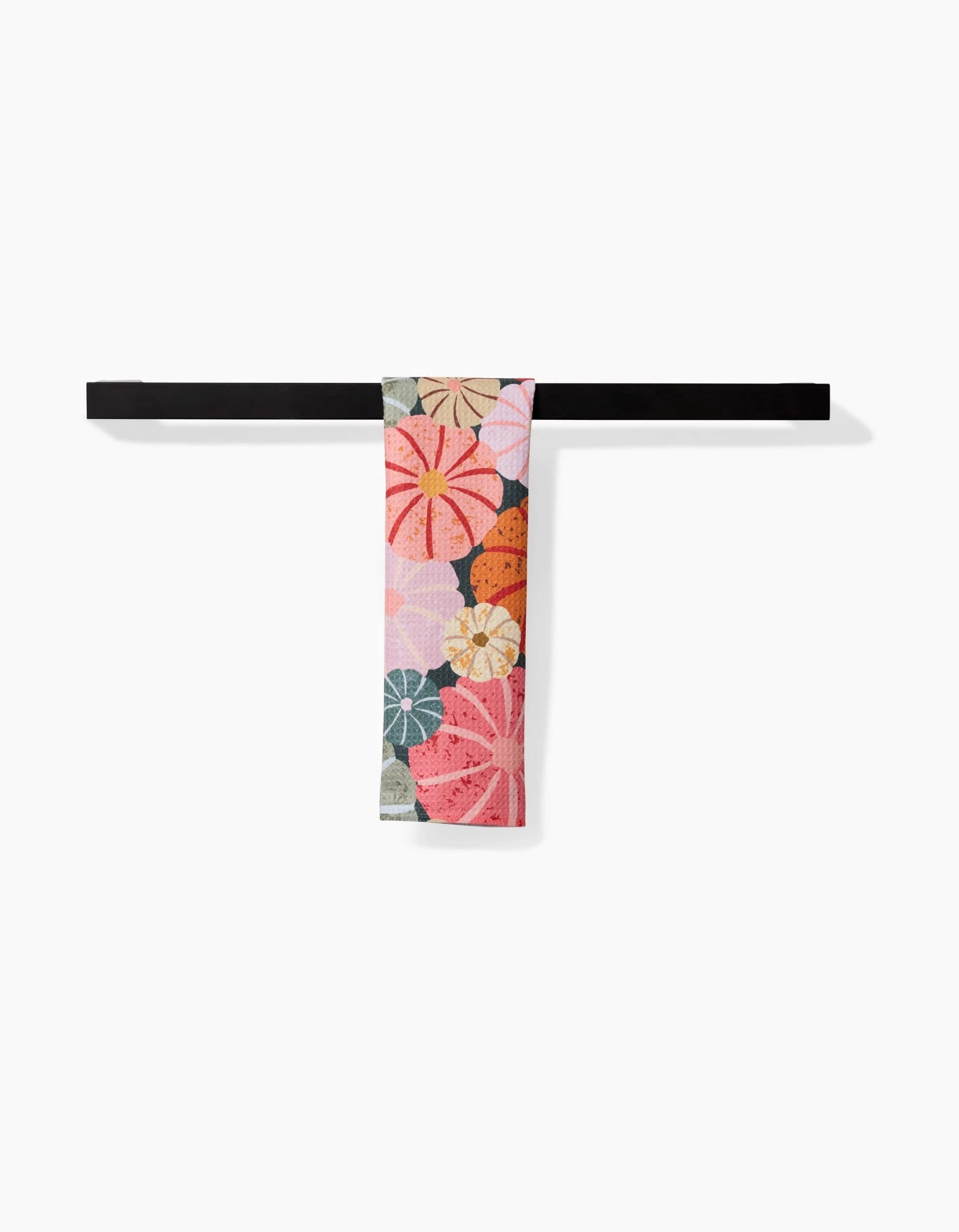 The Gorgeous Gourds towel by Rebecca Bobko, featuring a bold floral pattern in pink, orange, red, and green, hangs neatly folded over a sleek black bar against a white background.