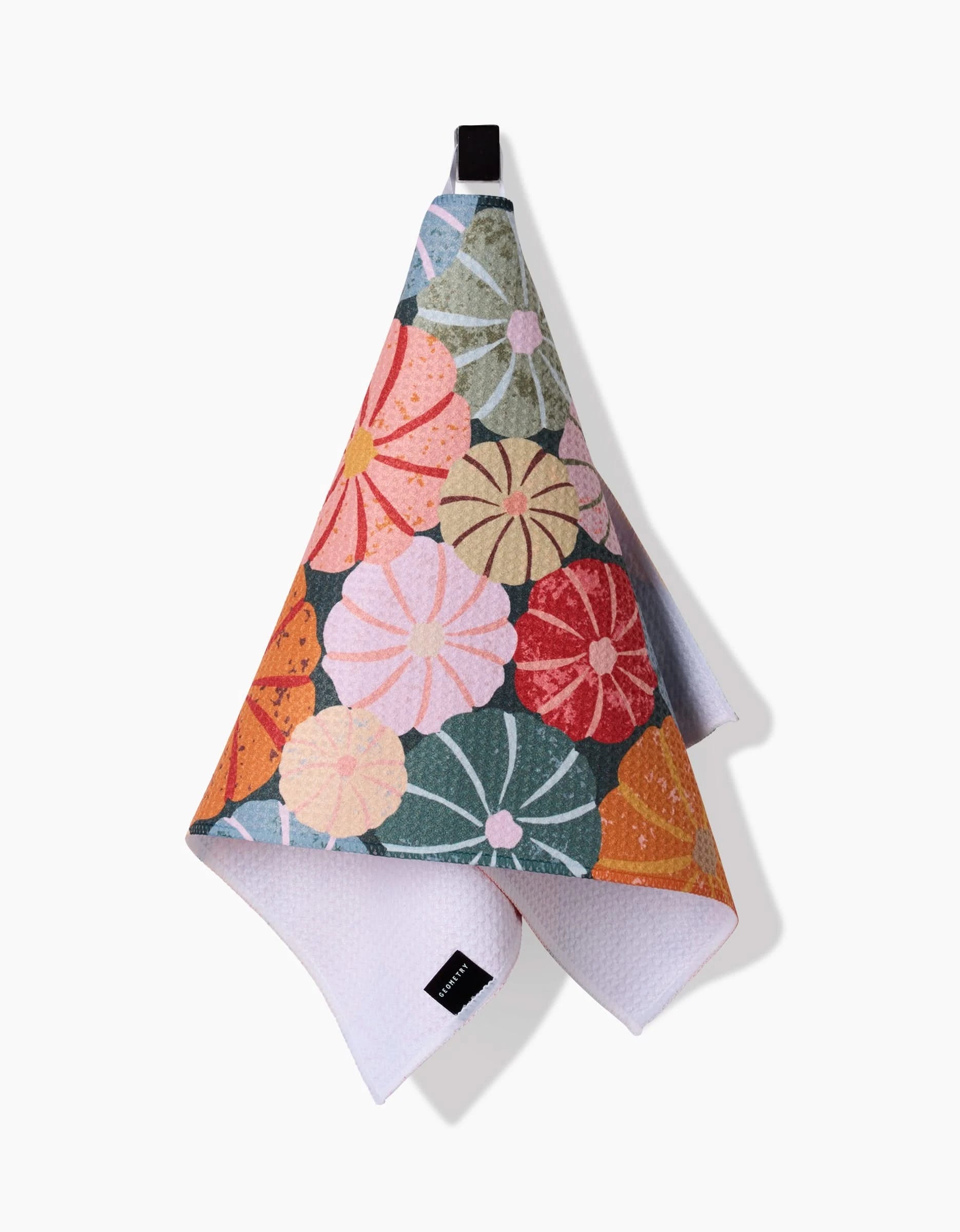 The Rebecca Bobko Gorgeous Gourds towel is ultra-absorbent, featuring a vibrant pattern of large, overlapping flowers in pink, orange, green, and yellow. It hangs from a black loop; the reverse side is plain white with a textured finish.