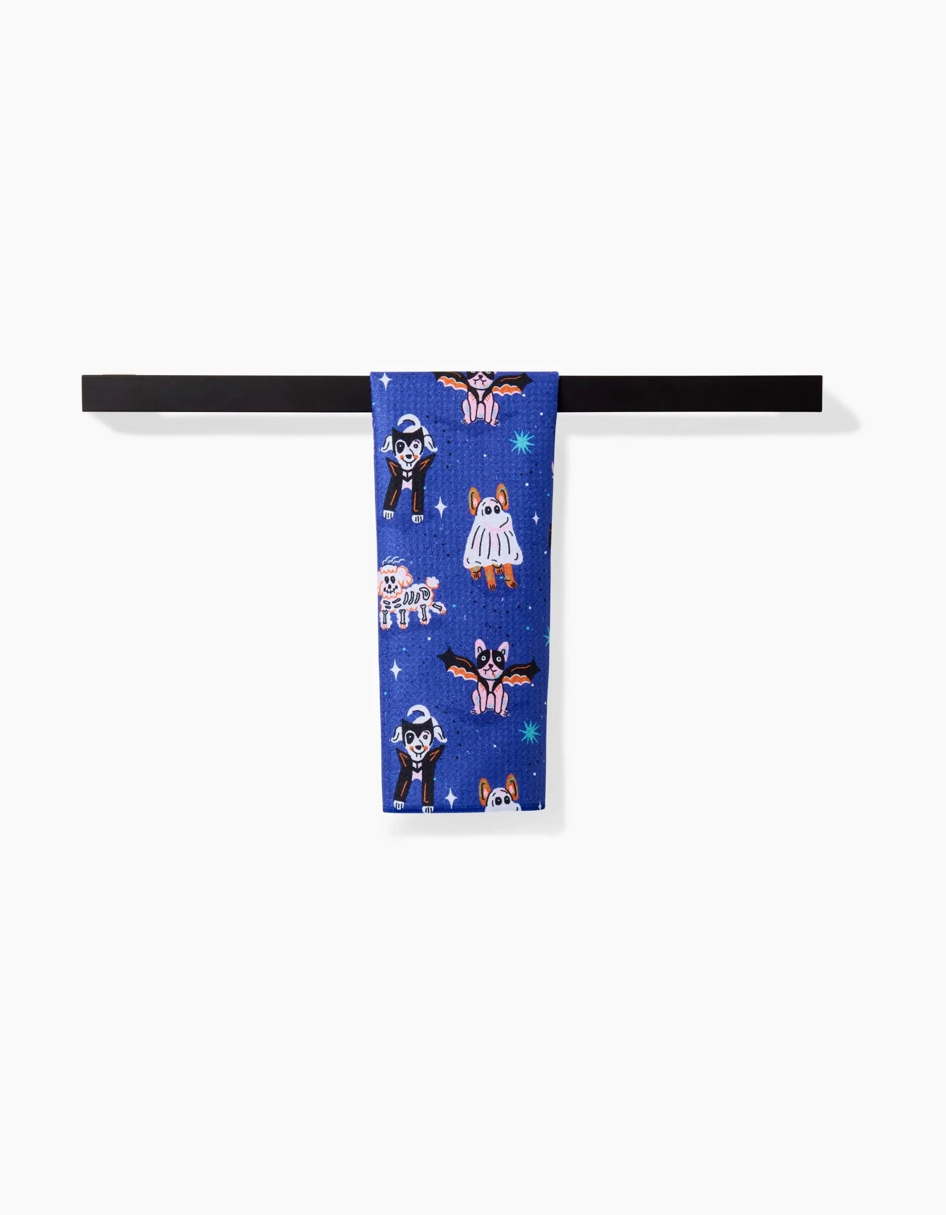 A folded blue “Halloween Hounds” Bar Towel by Camila Leão, featuring colorful cartoon bats, cats, and skeletons on soft Geoweave™ fabric, hangs over a black bar against a white background.