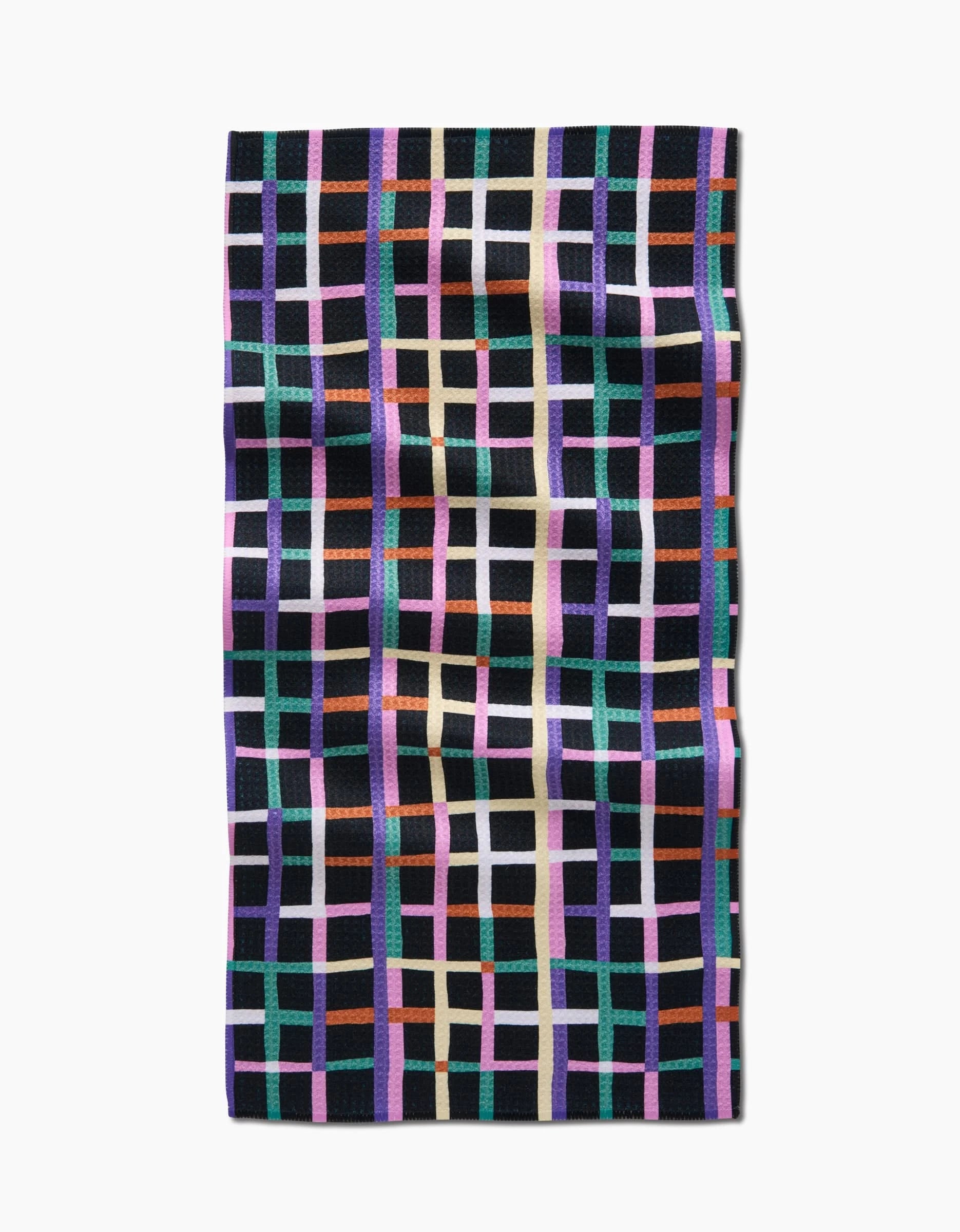 The Claire Iglesias Happy Plaid Bar Towel is crafted from Geoweave fabric and showcases a grid of intersecting purple, orange, green, beige, and black lines on a dark background. The towel is displayed flat with slight wrinkles.