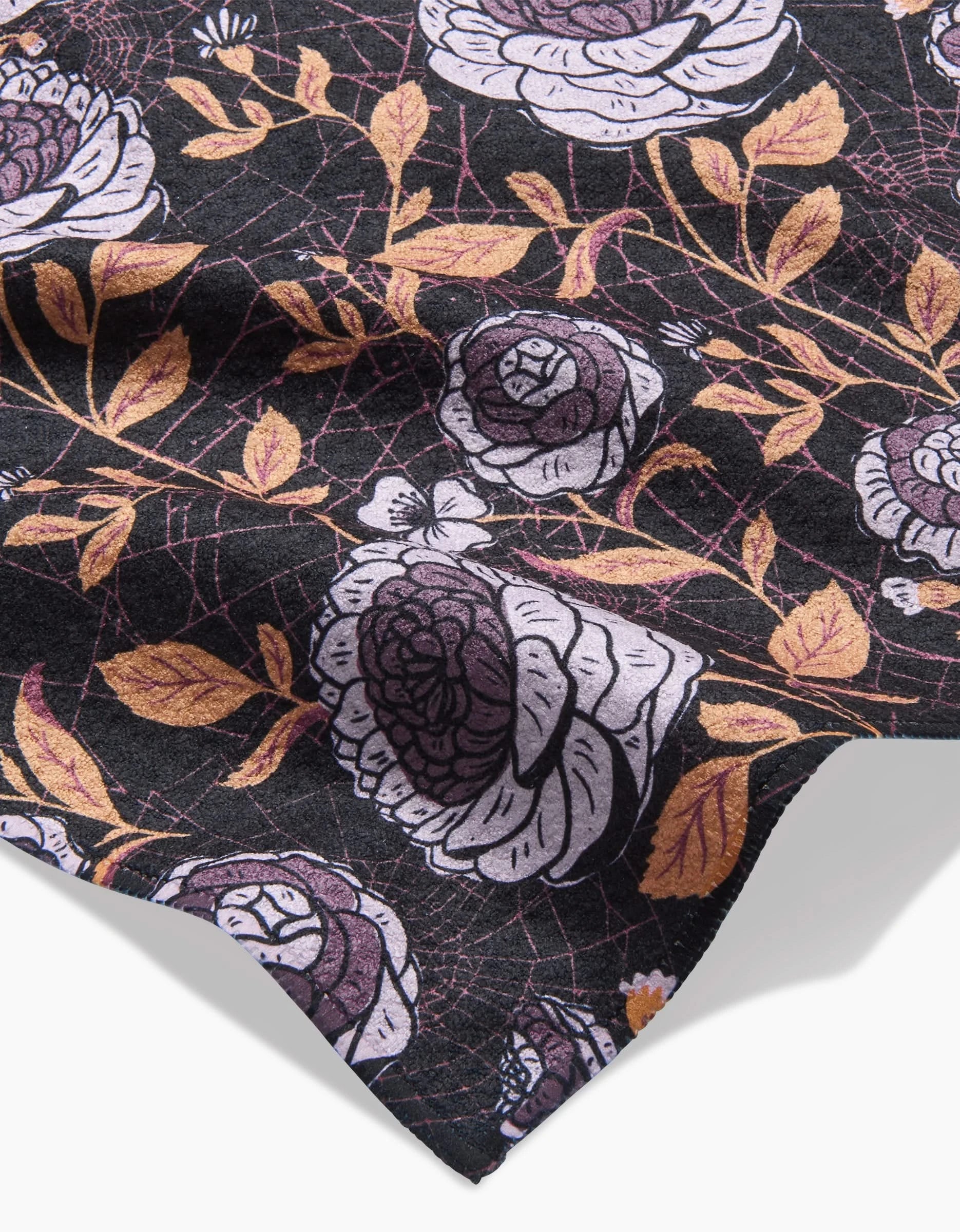 A close-up of the Haunted Roses towel by Danelys Sidron, featuring an ultra-absorbent fabric with white and purple roses and gold leaves on a black background. Gentle wrinkles add shadows and highlights to the floral design.