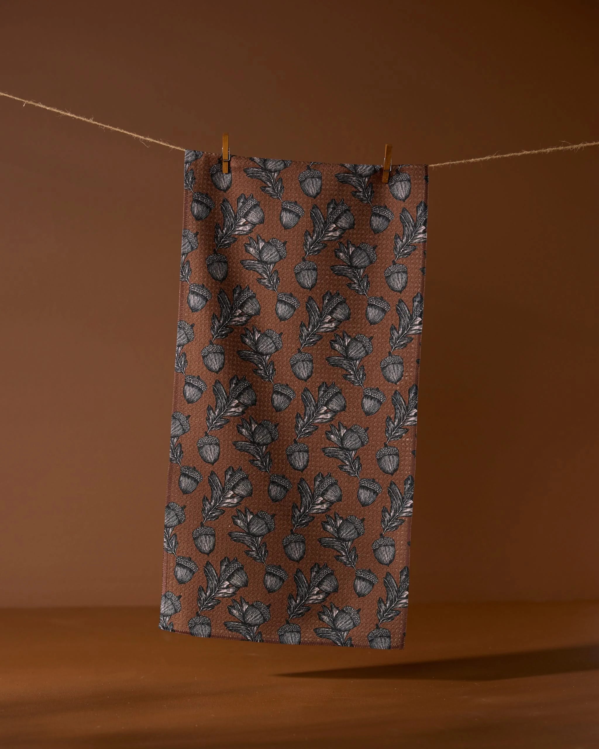 The Geometry Kindred towel, made from recycled materials, features a brown cloth with a repeating black leaf and acorn pattern. It hangs from a string with two clothespins against a brown background.