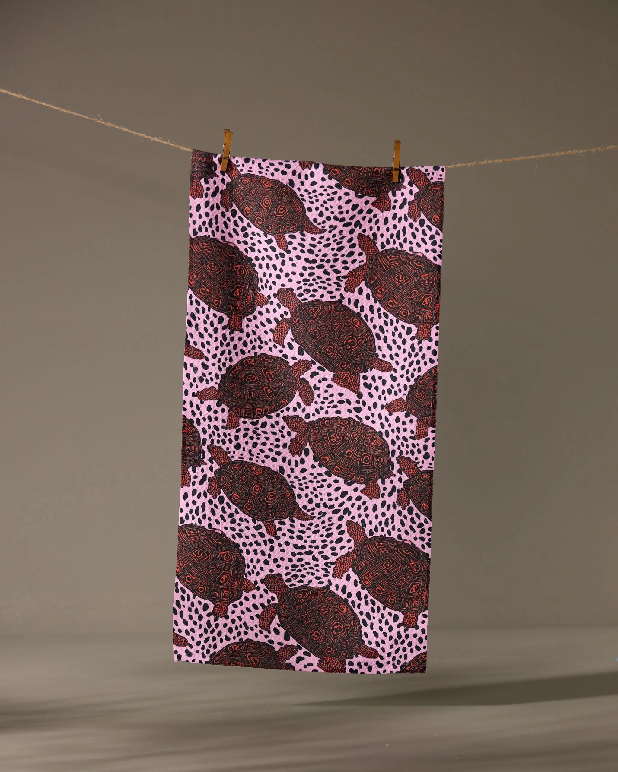 The Lagoon Crawl bar towel by Geometry features a pink geoweave fabric with a repeating black and red turtle pattern and black dots, hanging on a clothesline against a neutral background.