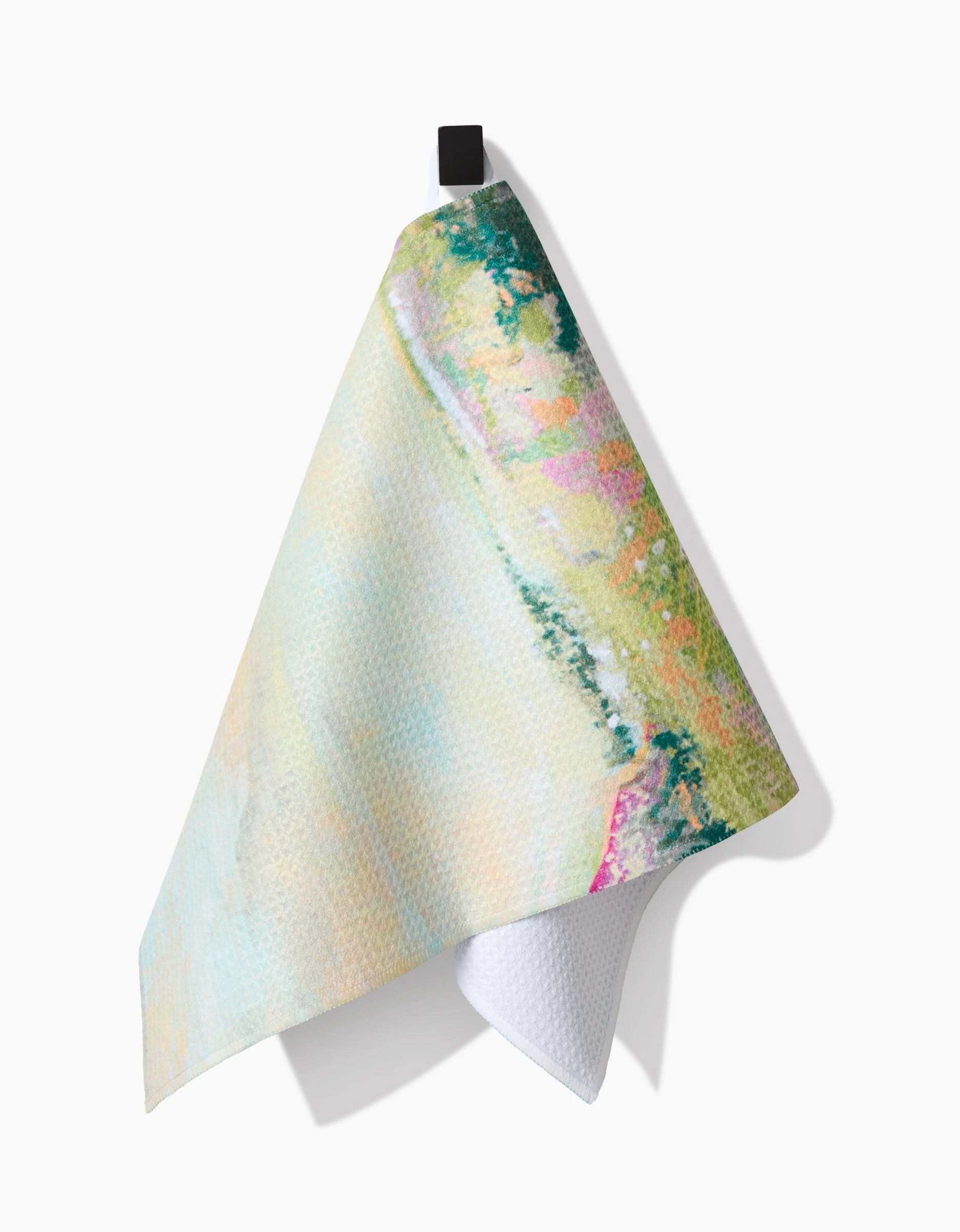 The Taryn Brennan Land of Milk and Honey bar towel features a pastel abstract watercolor pattern in green, yellow, and pink, made with eco-friendly recycled materials. It’s shown hanging from a black hook against a white background.